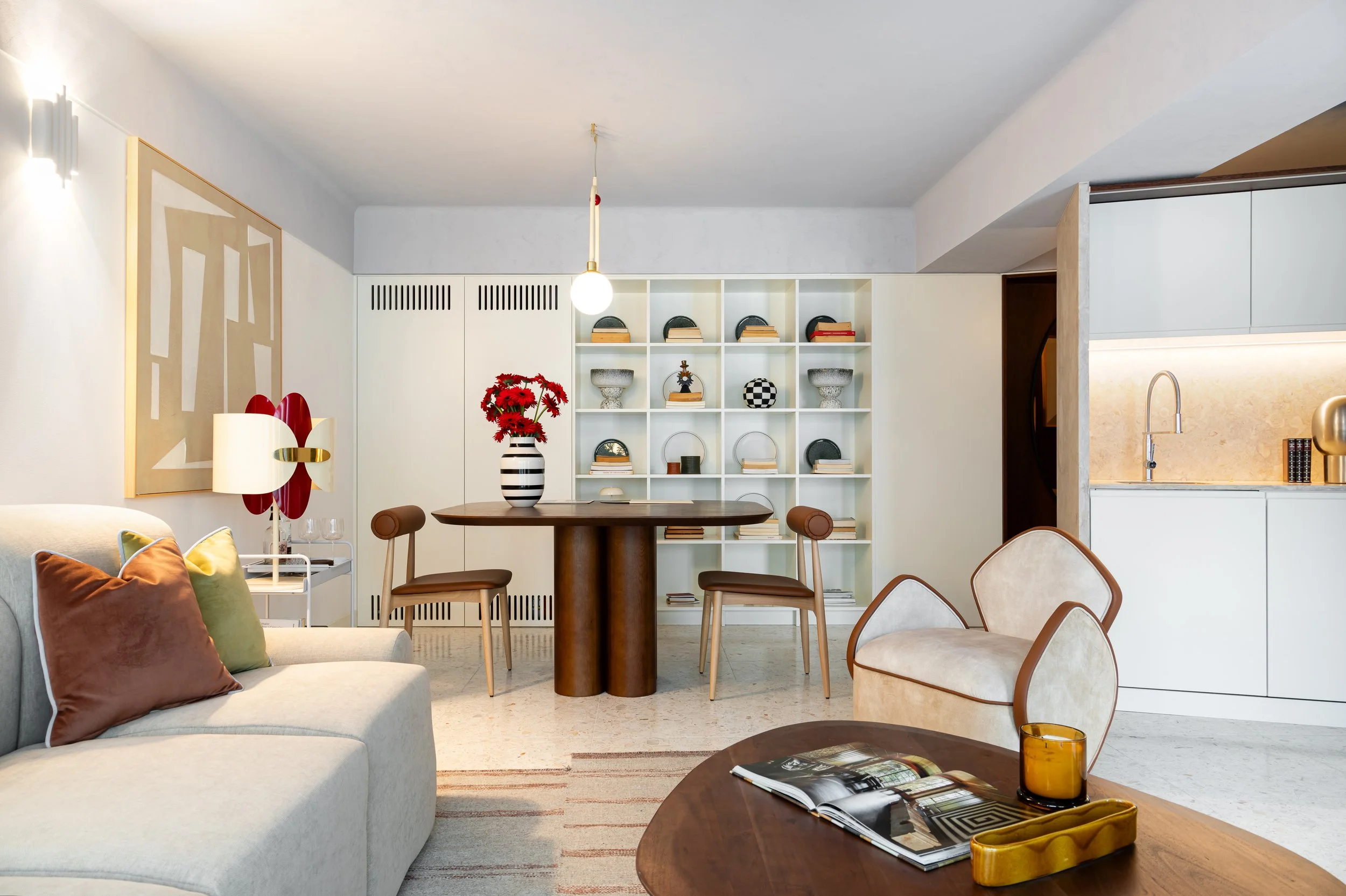 Living room with a sofa, decorative pillows, a coffee table with a magazine and a candle, a dining table with chairs and a vase with red flowers, a white shelving unit with decorative objects, and part of a kitchen with a sink.