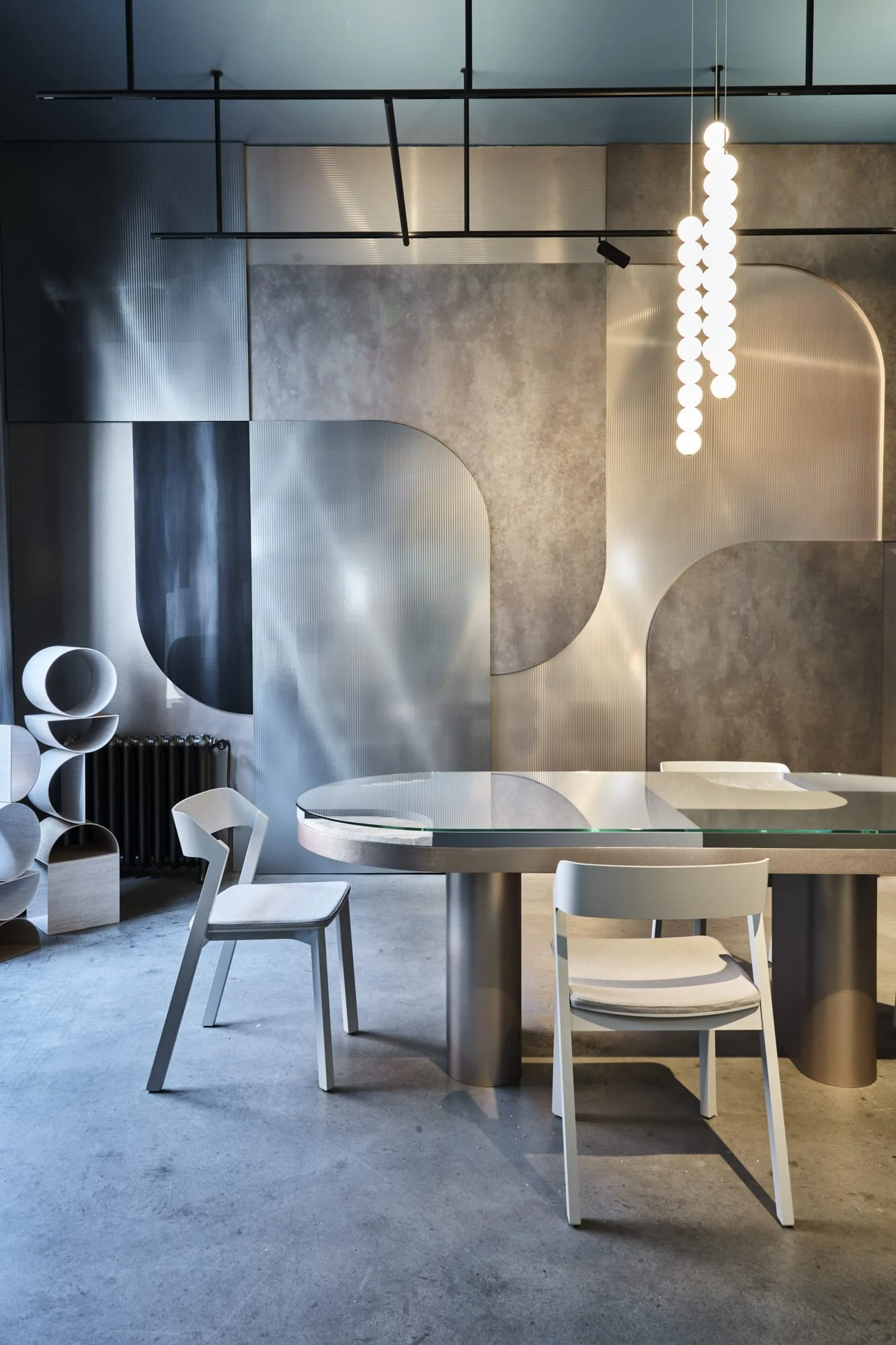 Modern minimalist interior with a round glass table, two white chairs, and abstract geometric wall panels with a lighting fixture hanging from the ceiling.