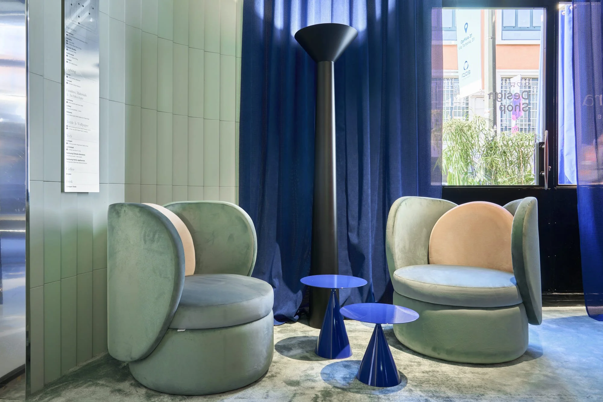 Two plush, vintage-style armchairs in pastel green with rounded backs and armrests, separated by two small, matching blue side tables with conical bases, positioned in front of a window with blue curtains, and a tall black floor lamp between the chairs.