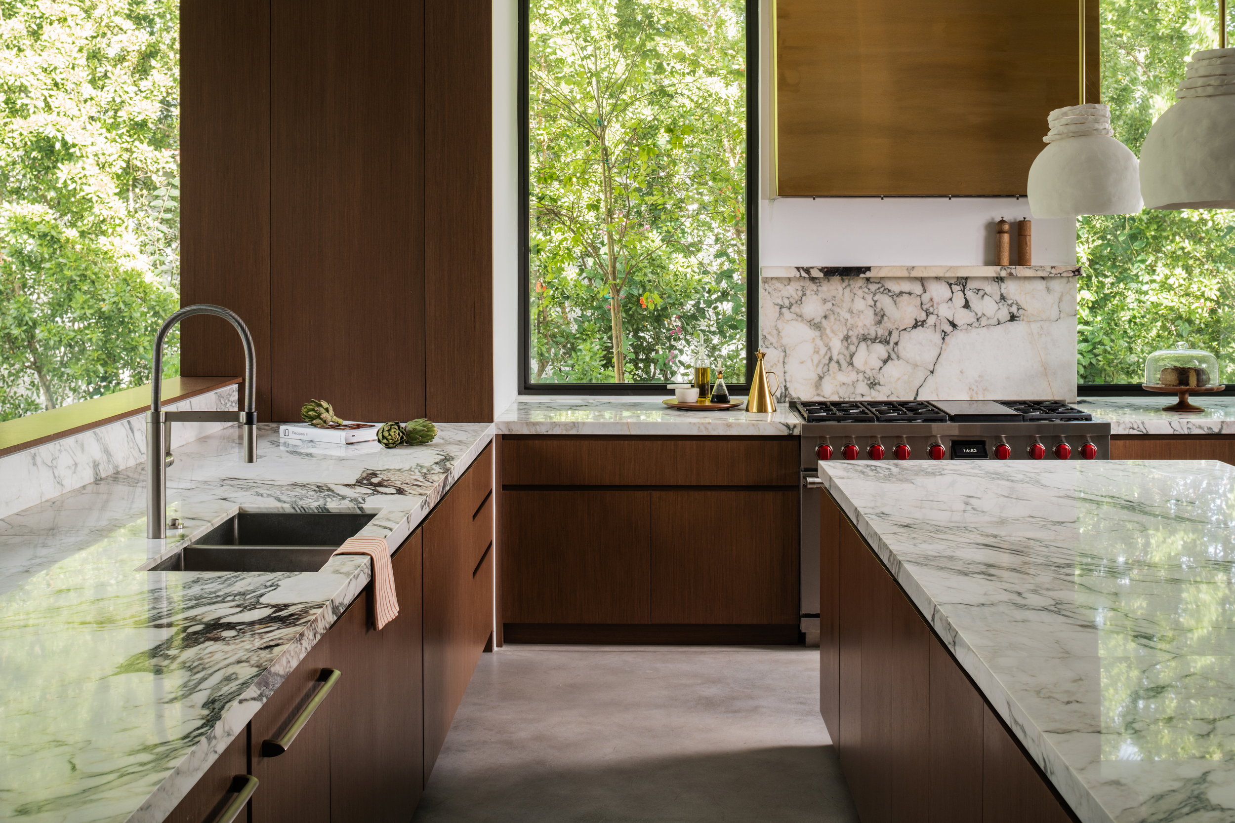 Modern kitchen with dark wood cabinets, marble countertops, a stainless steel sink, a large window overlooking green trees, and a stainless steel stove with red knobs.