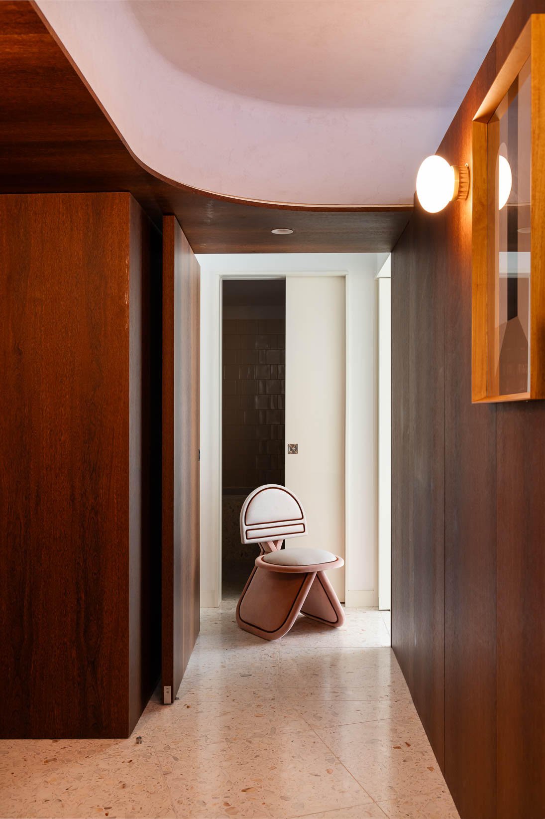 A modern, minimalist hallway with wooden wall and ceiling panels, a light-colored floor, and a uniquely designed pink chair with a round padded seat and curved backrest placed against a white door at the end of the hall. The lighting includes fixtures on the walls.