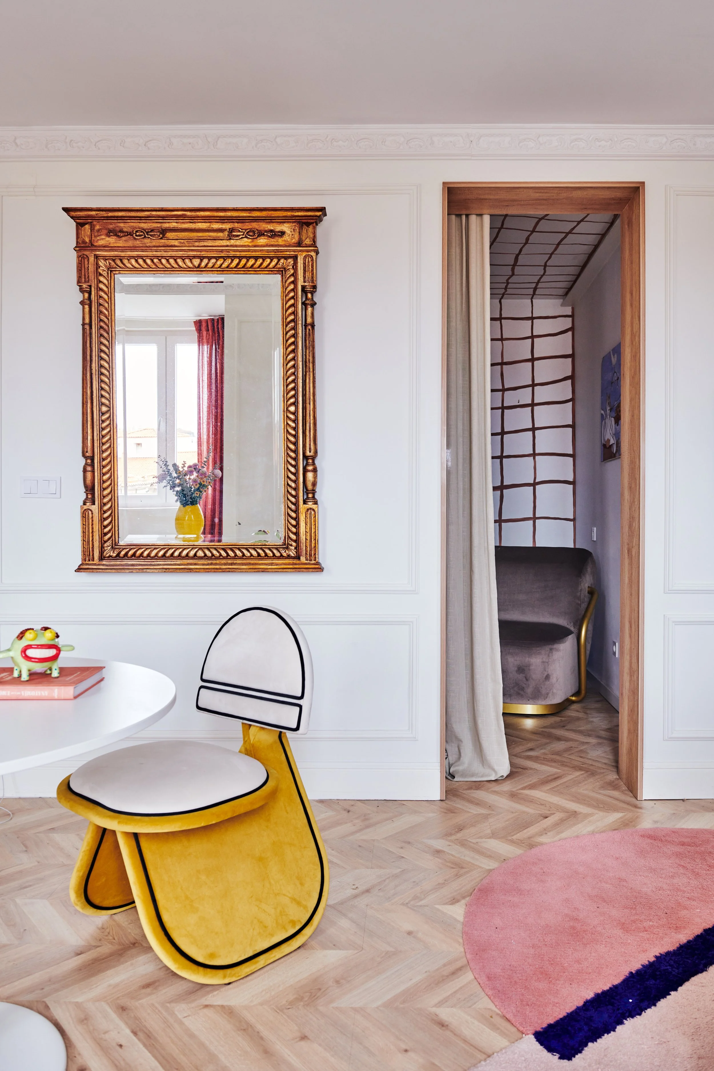 Interior of a living room with a white wall, large mirror with a gold ornate frame, a yellow and white chair at a white table, a pink and navy rug, and an opening to another room with a curtain and a gray sofa.