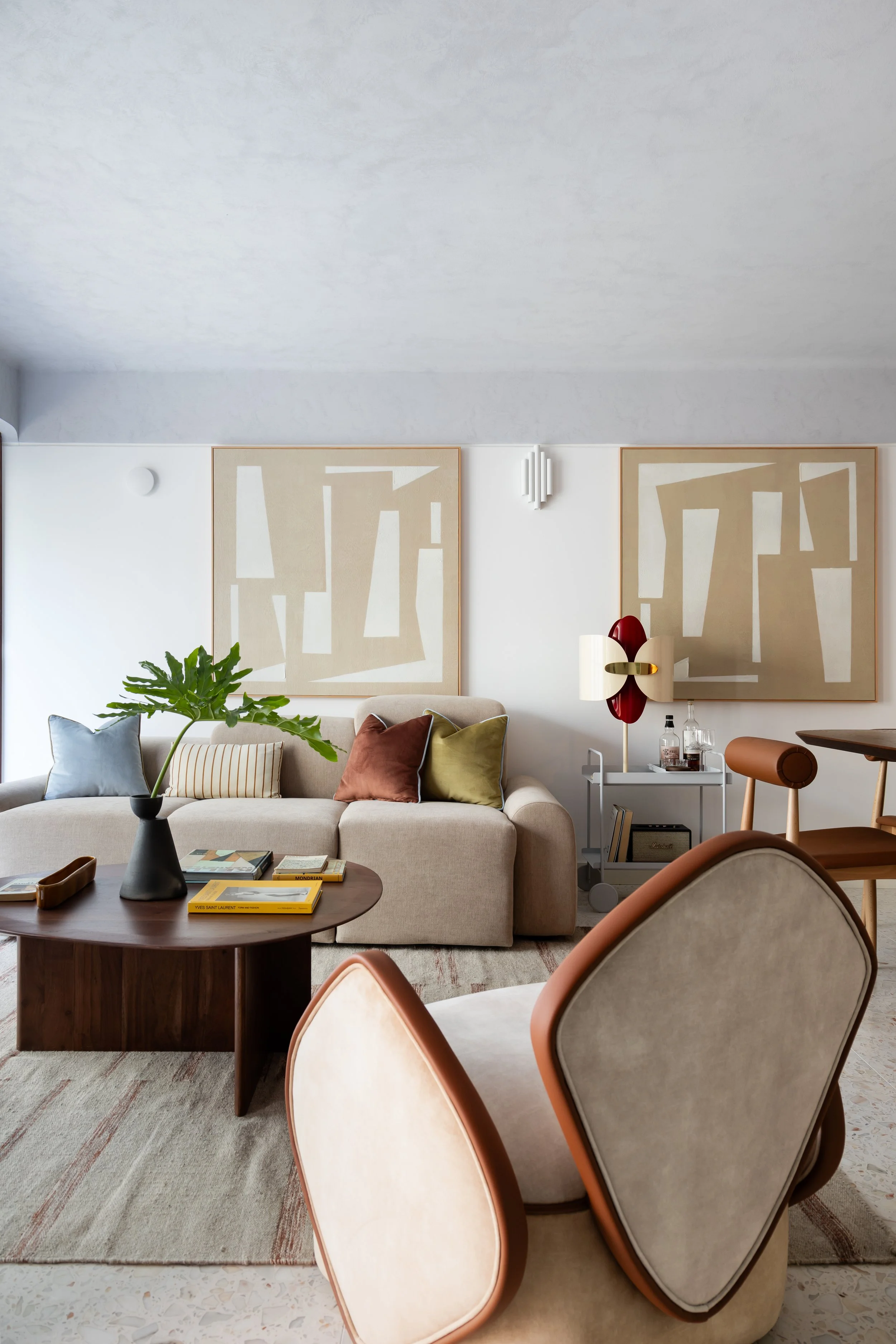 Living room with beige sofa, colorful pillows, wooden coffee table with books, large green plant in black vase, abstract wall art, side table with lamp, and two cream-colored chairs with brown trim.