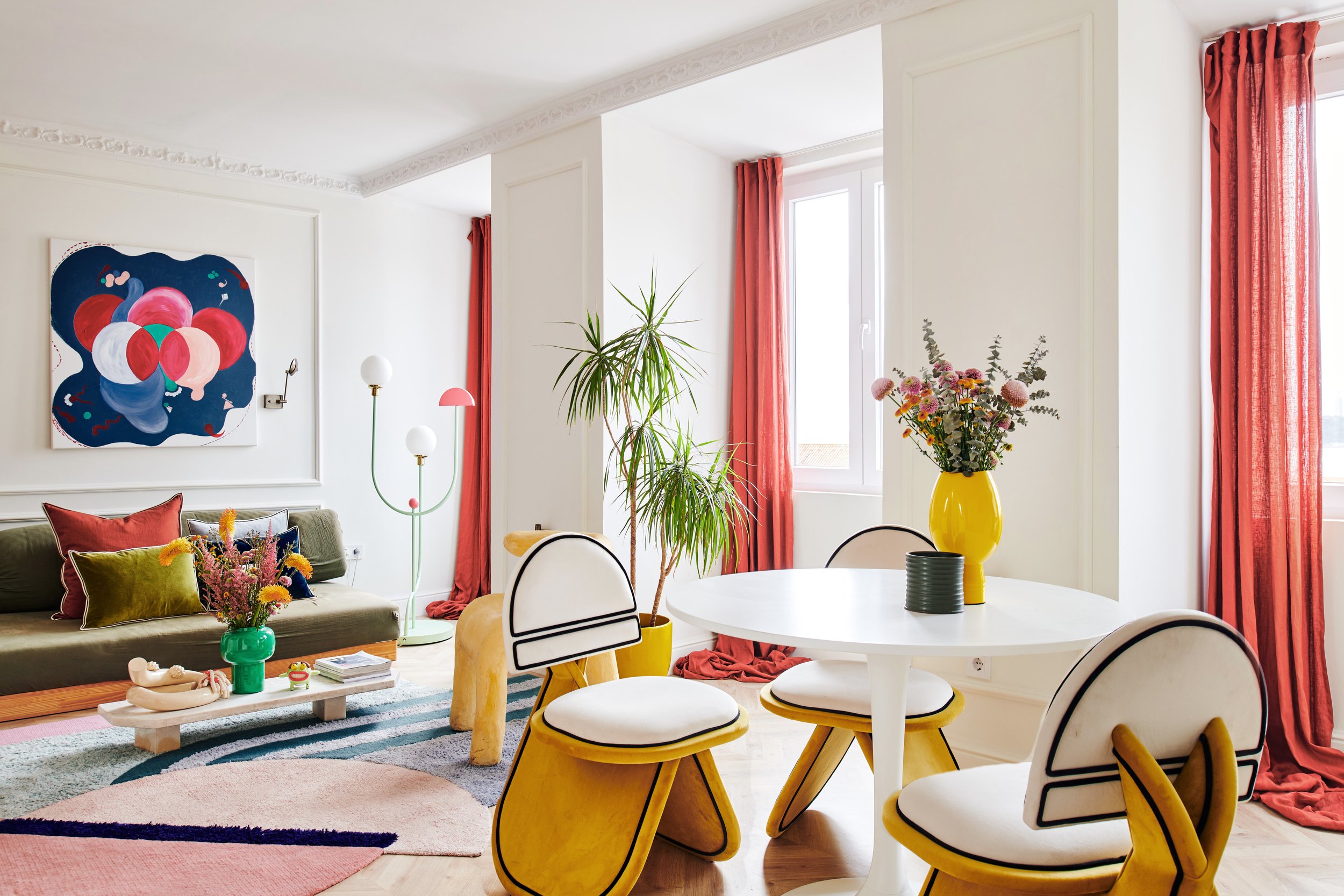 Living room with white walls, red curtains, a white round table with four yellow and white chairs, a green sofa with colorful pillows, a coffee table with a green vase and flowers, an abstract artwork on the wall, a multicolored rug, a yellow vase with pink and purple flowers, and a large potted plant.