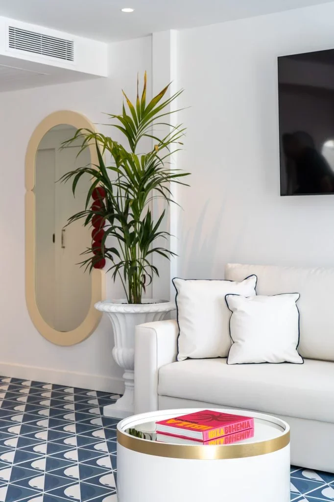 Modern living room with white sofa, white pillows with navy piping, a round white coffee table with gold trim, a tall potted plant in a decorative white planter, a mirror with a beige border, and a large flat-screen TV on the wall, with patterned black and white tiled flooring.