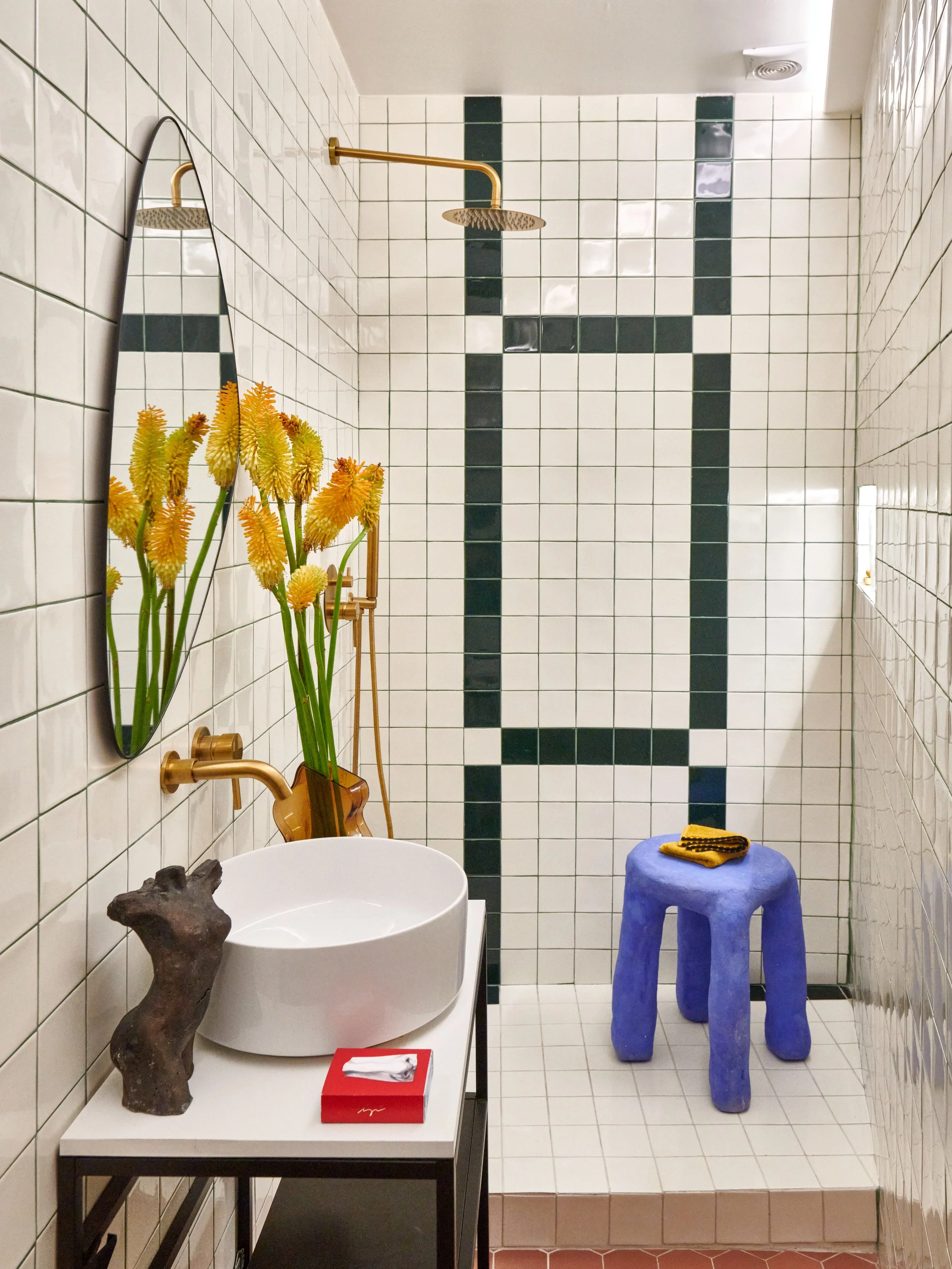 Bathroom shower area with white tiles accented by dark green tiles outlining a rectangle pattern, blue stool with folded towel on top, round white vessel sink on a black metal stand with decorative items including a branch, a mirror reflecting yellow flowers, and gold showerhead.