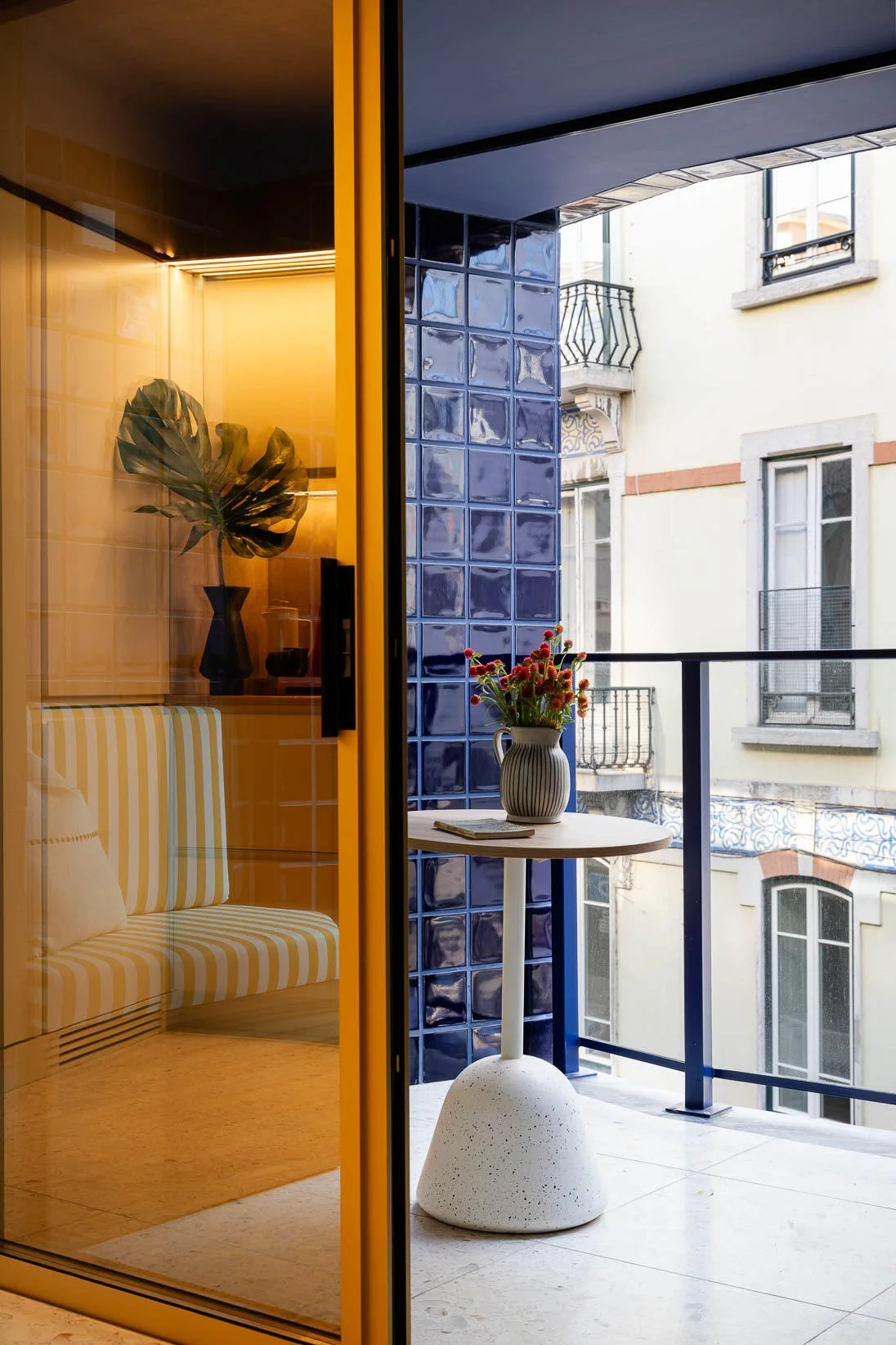 A small balcony with a table holding a vase of flowers, a building with decorative balconies and windows in the background, and a glass door with a yellow frame revealing an interior with a striped cushion and indoor planter.