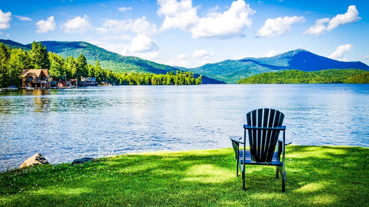A black Adirondack chair on a grassy lakeshore overlooking a calm lake, with green trees, houses on the water, and mountains in the background under a partly cloudy sky.