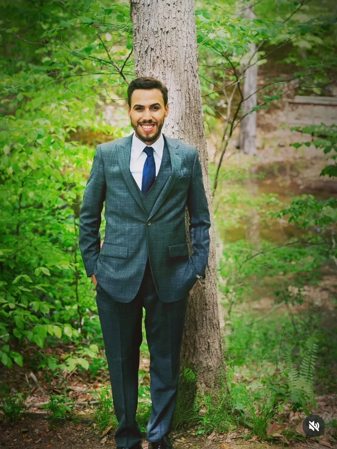 A man in a suit smiling and standing outdoors next to a tree in a green wooded area.