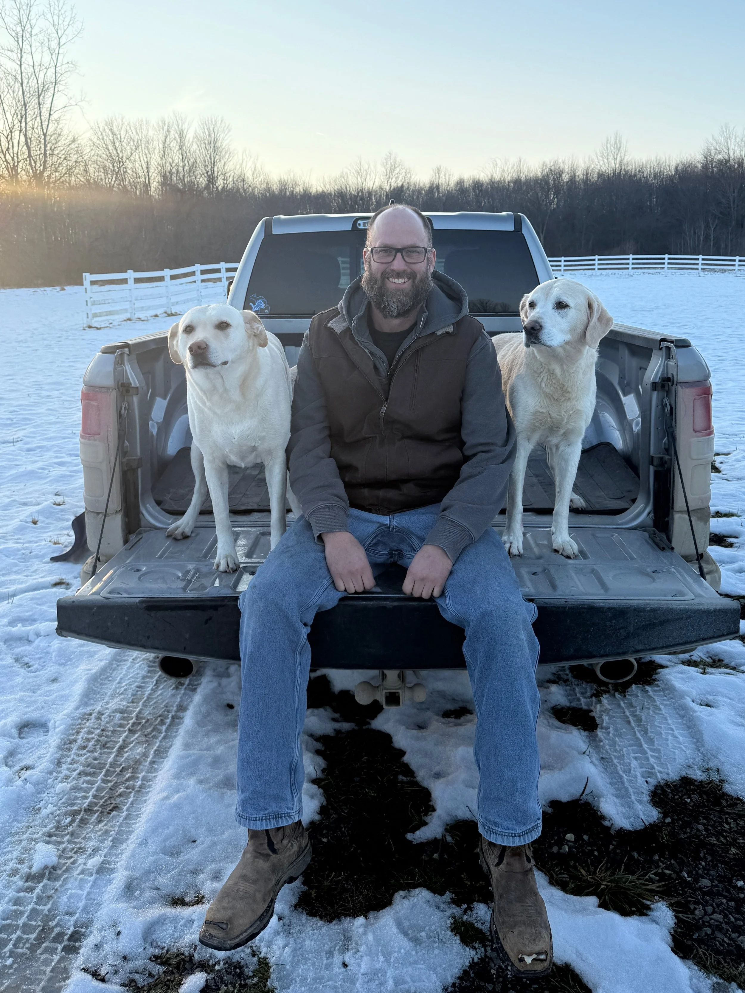 A man with glasses and a beard sitting on the tailgate of a pickup truck in a snowy field, flanked by two yellow Labrador retrievers, smiling at the camera during sunset.