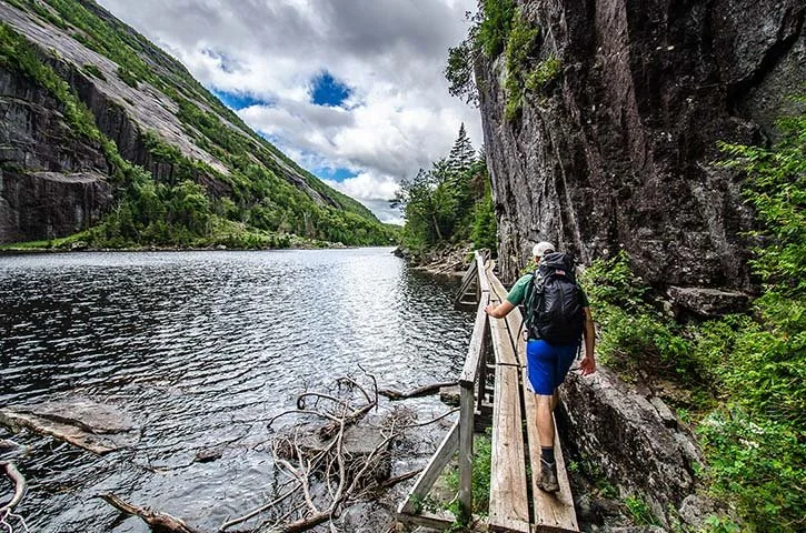 Hiker walking along a wood boardwalk against steep cliffs on the Champlain Adirondack Biosphere Avalanche Lake Trail.