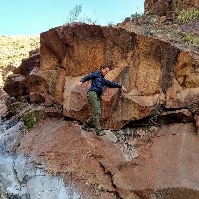 A woman standing on a large rock in a desert landscape, surrounded by reddish-brown rocks and sparse vegetation.