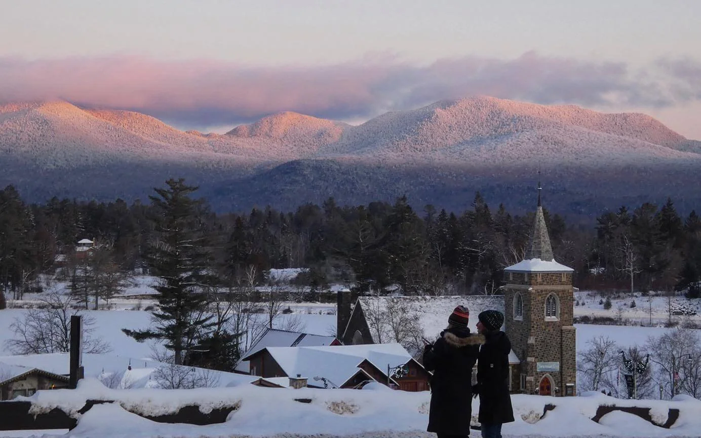 Two women in long coats looking over the snow covered town of Lake Placid with white mountains in the background in the Champlain-Adirondack Biosphere Network.