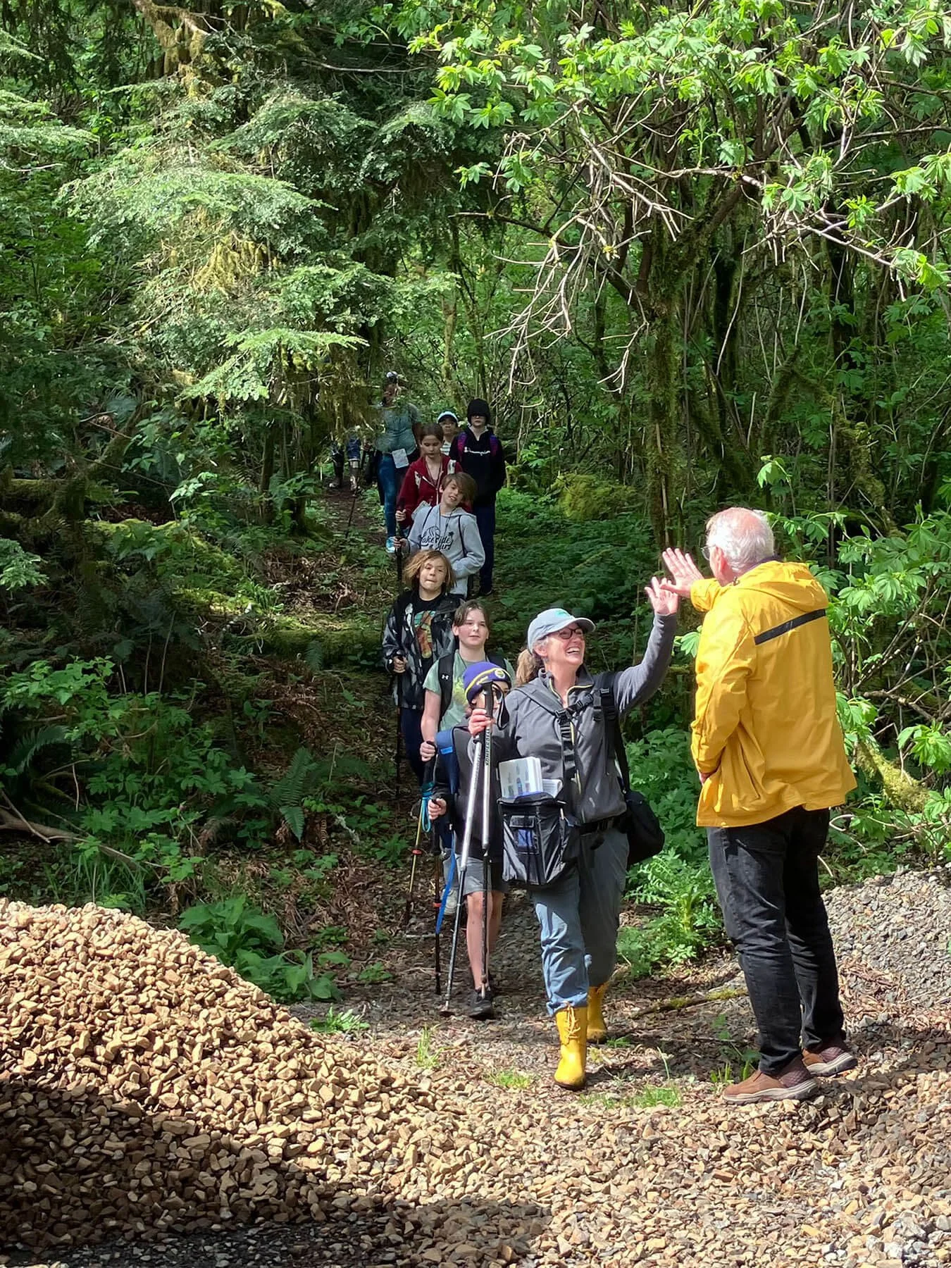 A woman leading a group of young hikers through a tree-filled trail in Oregon's Cascade Head Biosphere Region high fives an older man wearing a yellow rain coat.