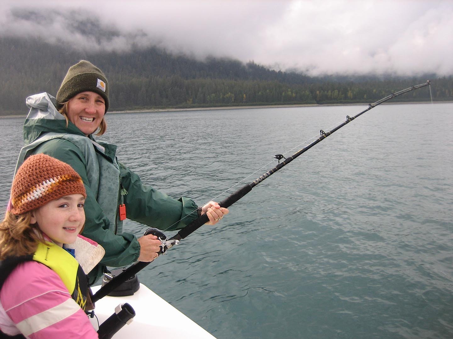 A woman and a young girl fishing from a boat on a lake with a mountainous, foggy background.