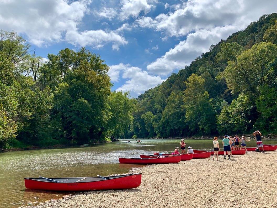 Row of red canoes with people getting into them along a calm river with full green trees in the Mammoth Cave Biosphere Region.
