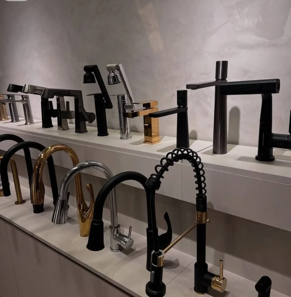 Estantería con diversas llaves de fregadero en diferentes estilos y acabados, incluyendo negro, dorado, plateado y combinaciones de estos, en una tienda de accesorios para cocina.