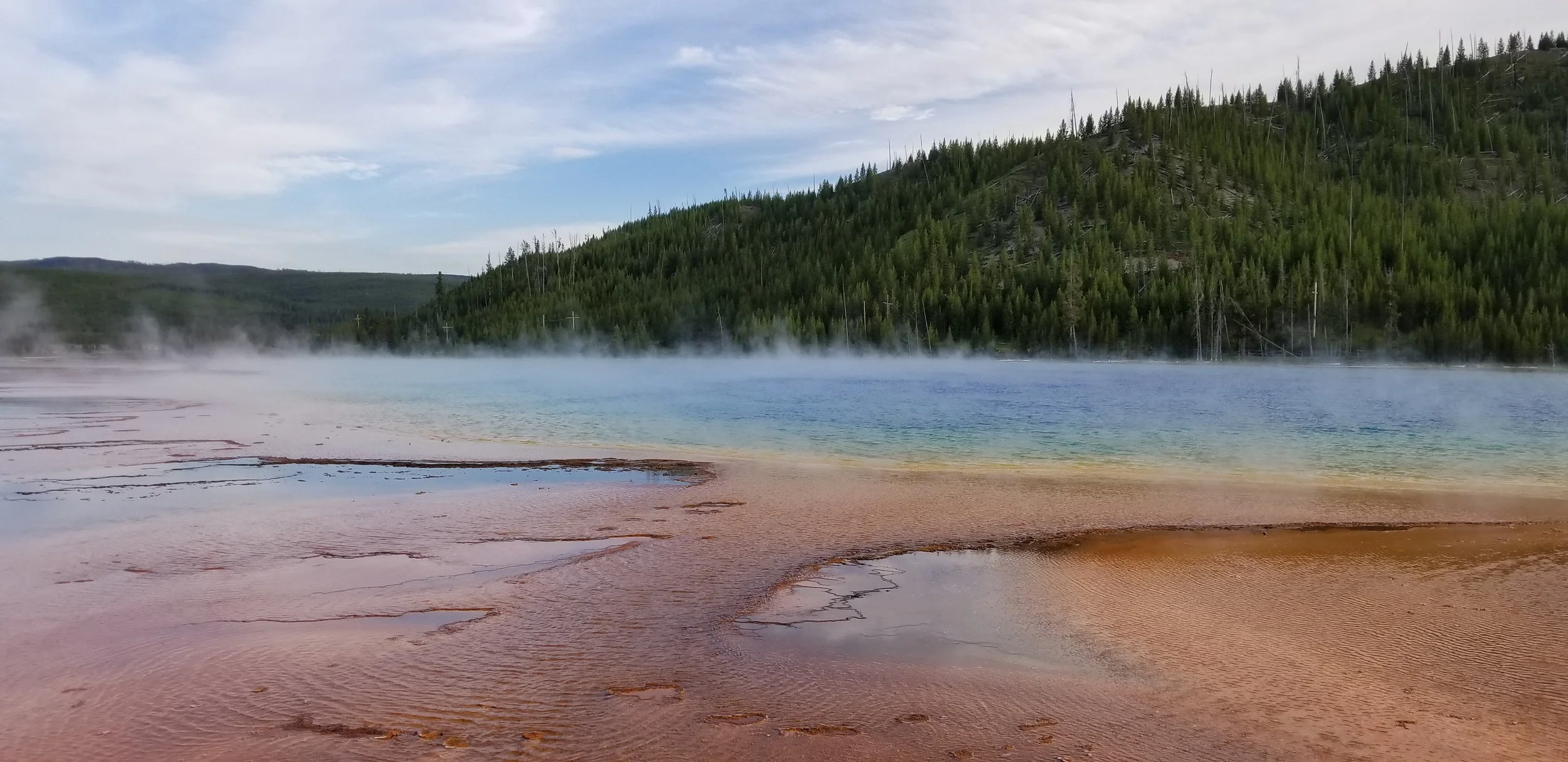 Photo of a geothermal hot spring with reddish-brown mineral deposits on the edge, steaming water, and a forested hillside in the background under a partly cloudy sky.