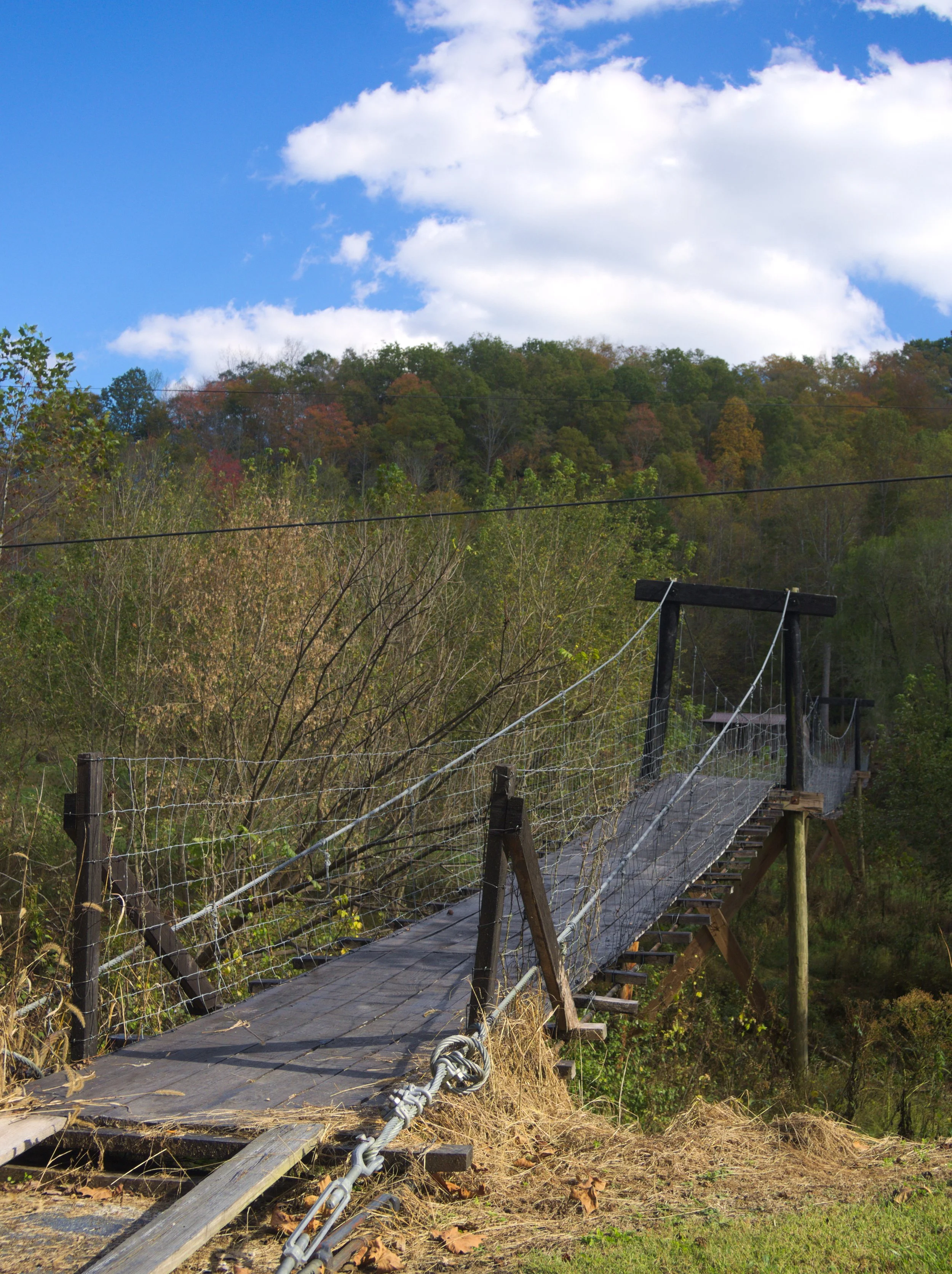 A wooden suspension bridge with safety netting, spanning a grassy and wooded area with hills and a partly cloudy sky above.