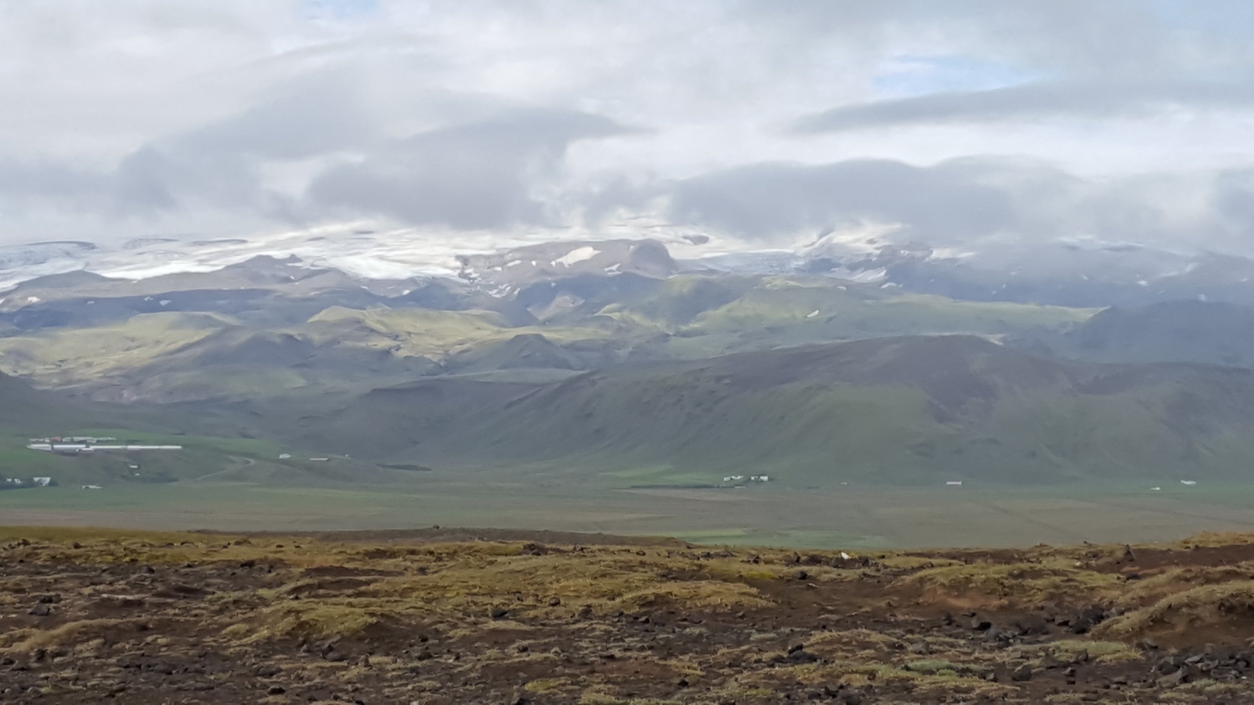 Scenic landscape of rolling green hills and mountains (Katla) with snow-capped peaks, under a cloudy sky.
