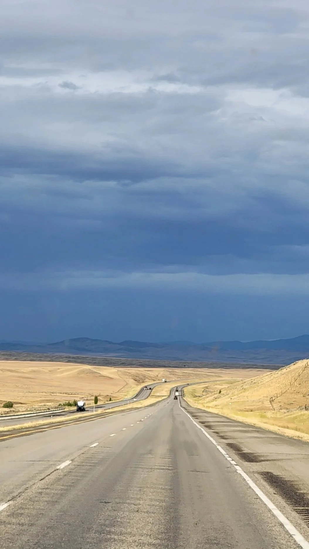 A long, winding road through a dry, open landscape with hills in the distance under a cloudy sky.