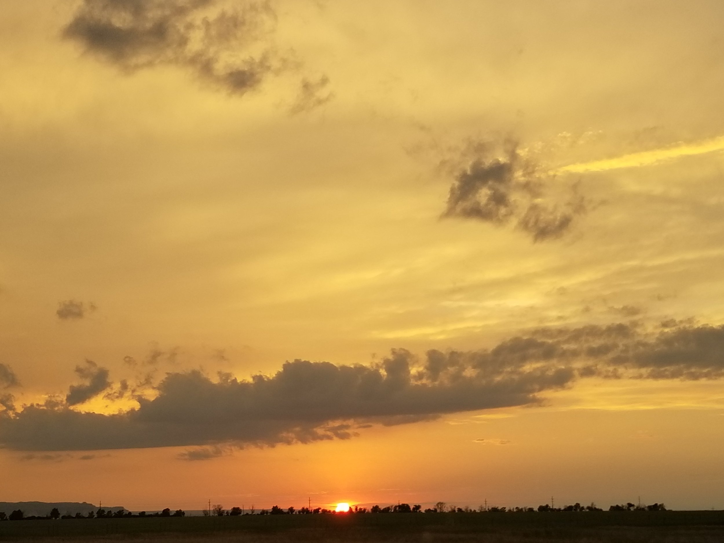 A sunset over a flat landscape with dark clouds in the sky and a horizon with some trees and power lines.