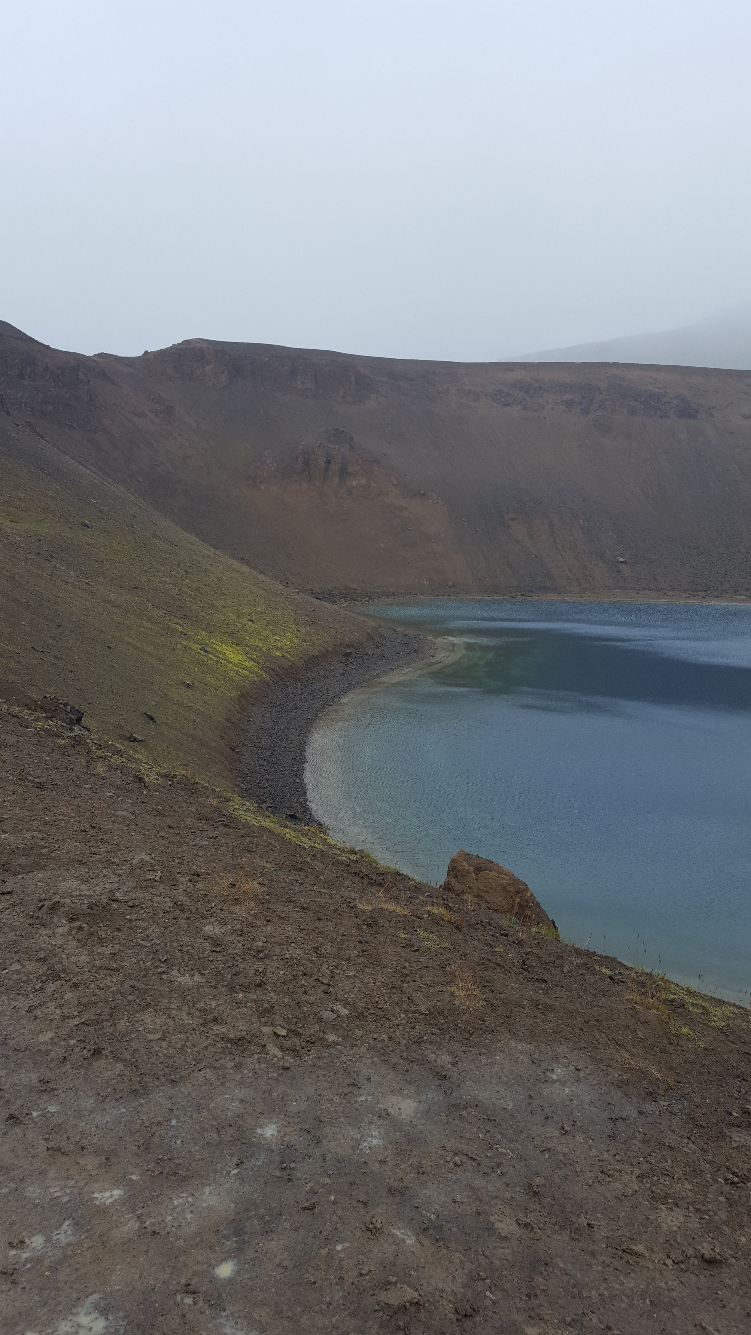 Víti Crater Lake in Krafla Caldera, with steep, rocky, and barren slopes surrounding it, under a cloudy sky.