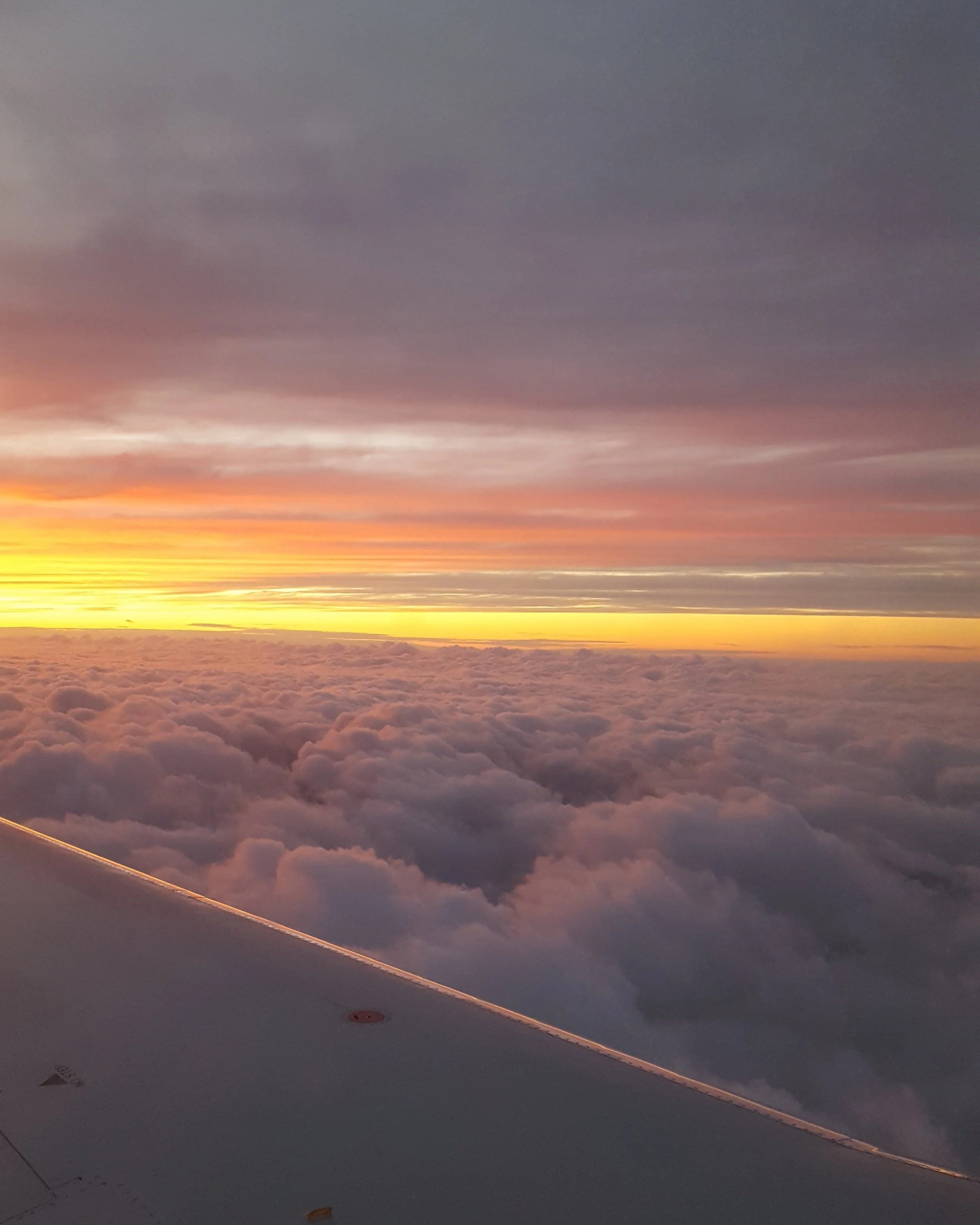 An organe and yellow sunset over lavender colored clouds, viewed over the wing of a plane.