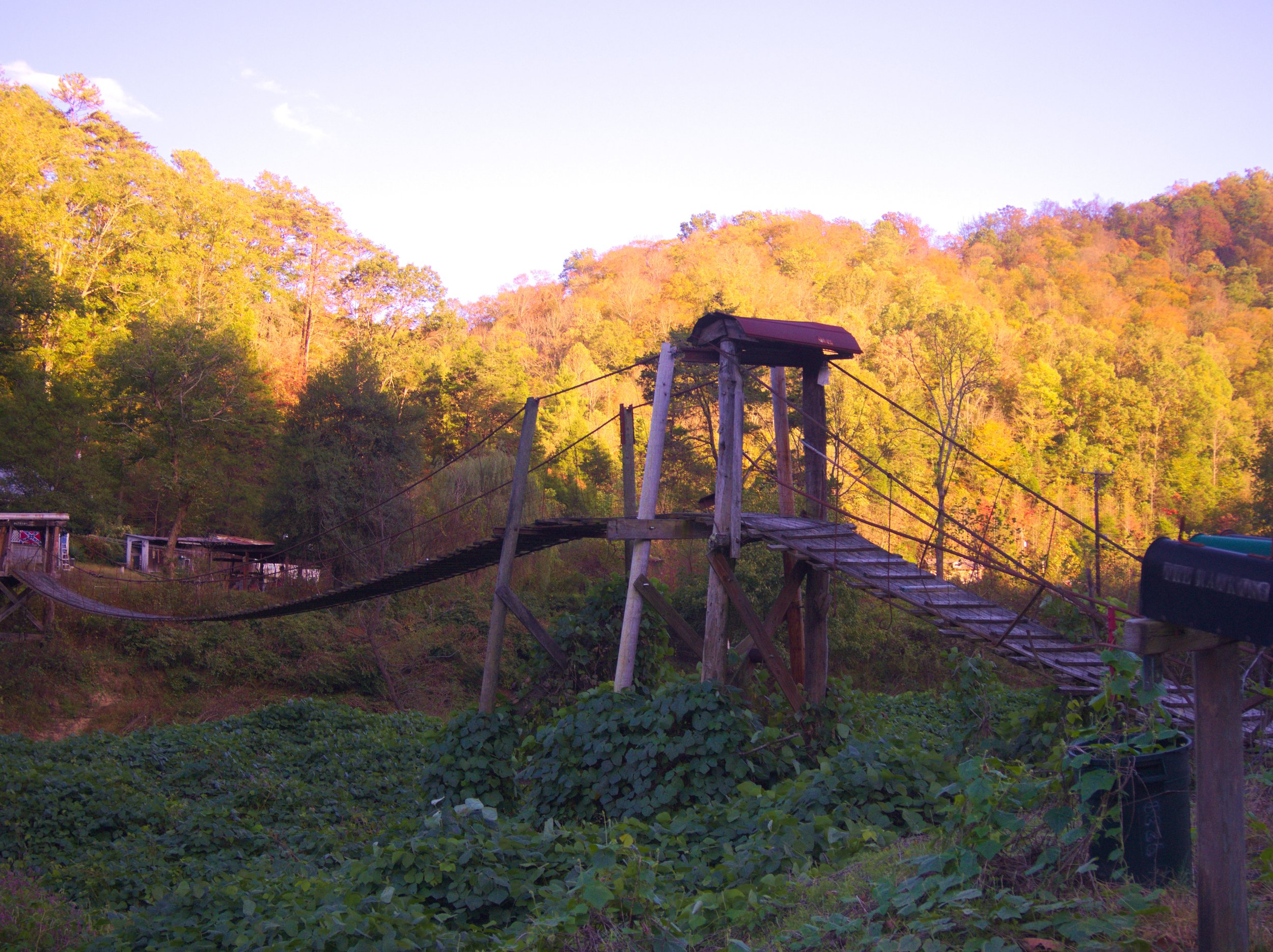 A rustic wooden suspension bridge in a rural area with green foliage, leading towards a hillside with trees in fall colors under a partly cloudy sky.