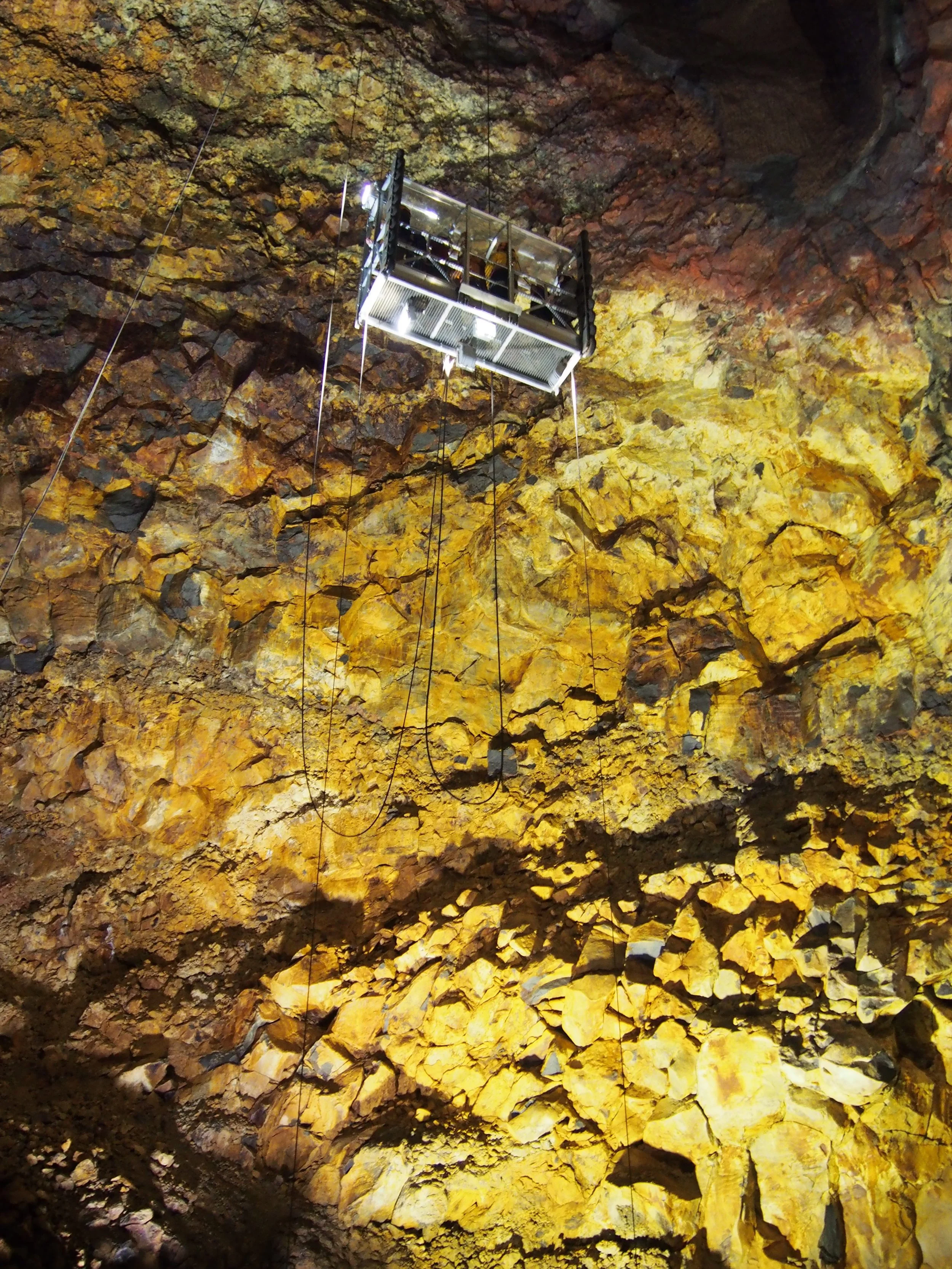 Inside a large cave (Þríhnúkagígur) with yellow and brown rock formations, a suspended metal platform or lift is hanging from the ceiling.