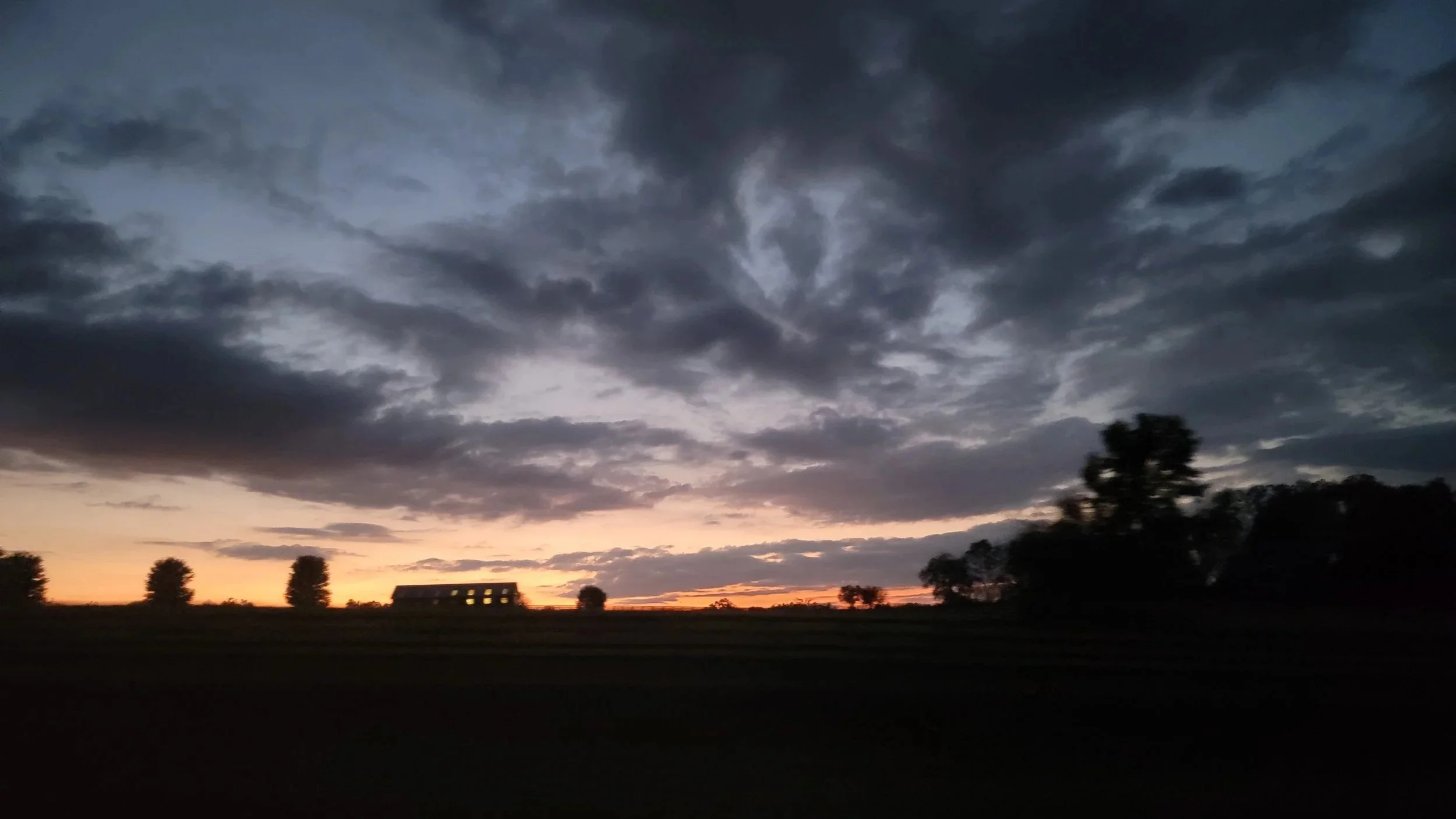 A landscape with a dramatic, cloudy sky during sunset; dark silhouettes of trees and a building with lit windows are visible on the horizon.