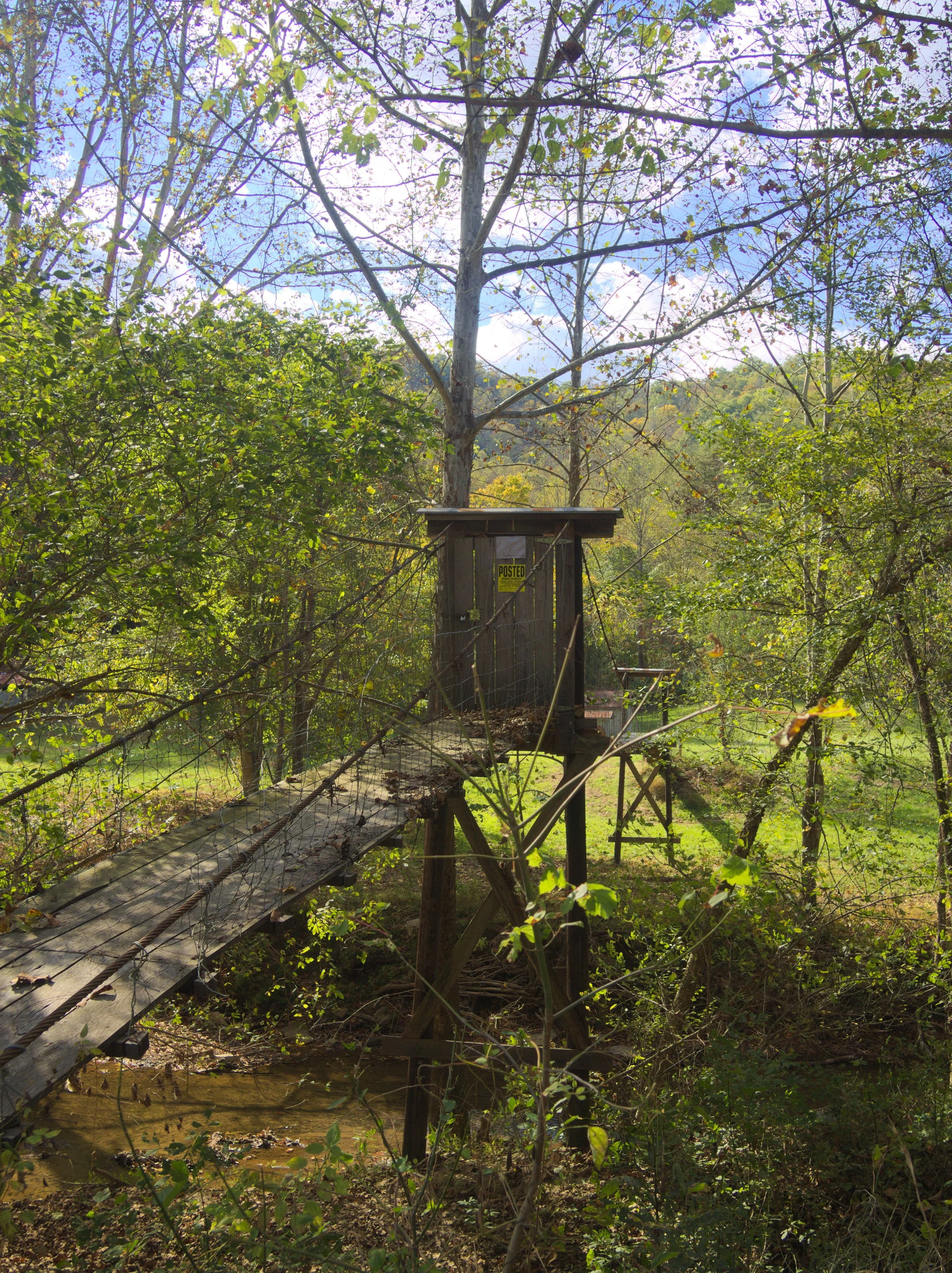 Wooden bridge with a ramp, located in a forested area with green trees and a partly cloudy sky.