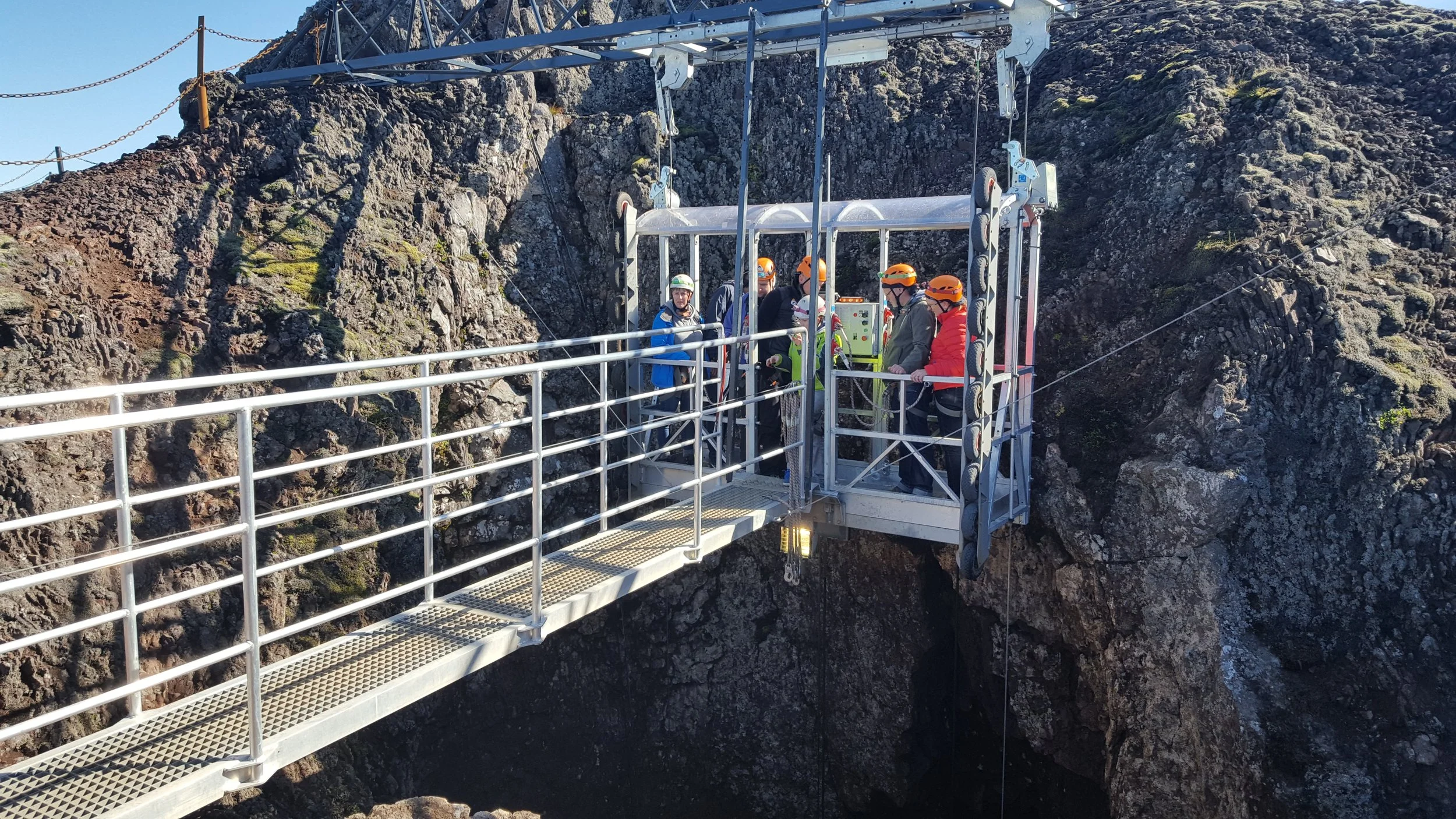 Group of people wearing helmets standing on an outdoor metal platform suspended between rocky cliffs (about to descend into Þríhnúkagígur), with some people looking at a control panel.