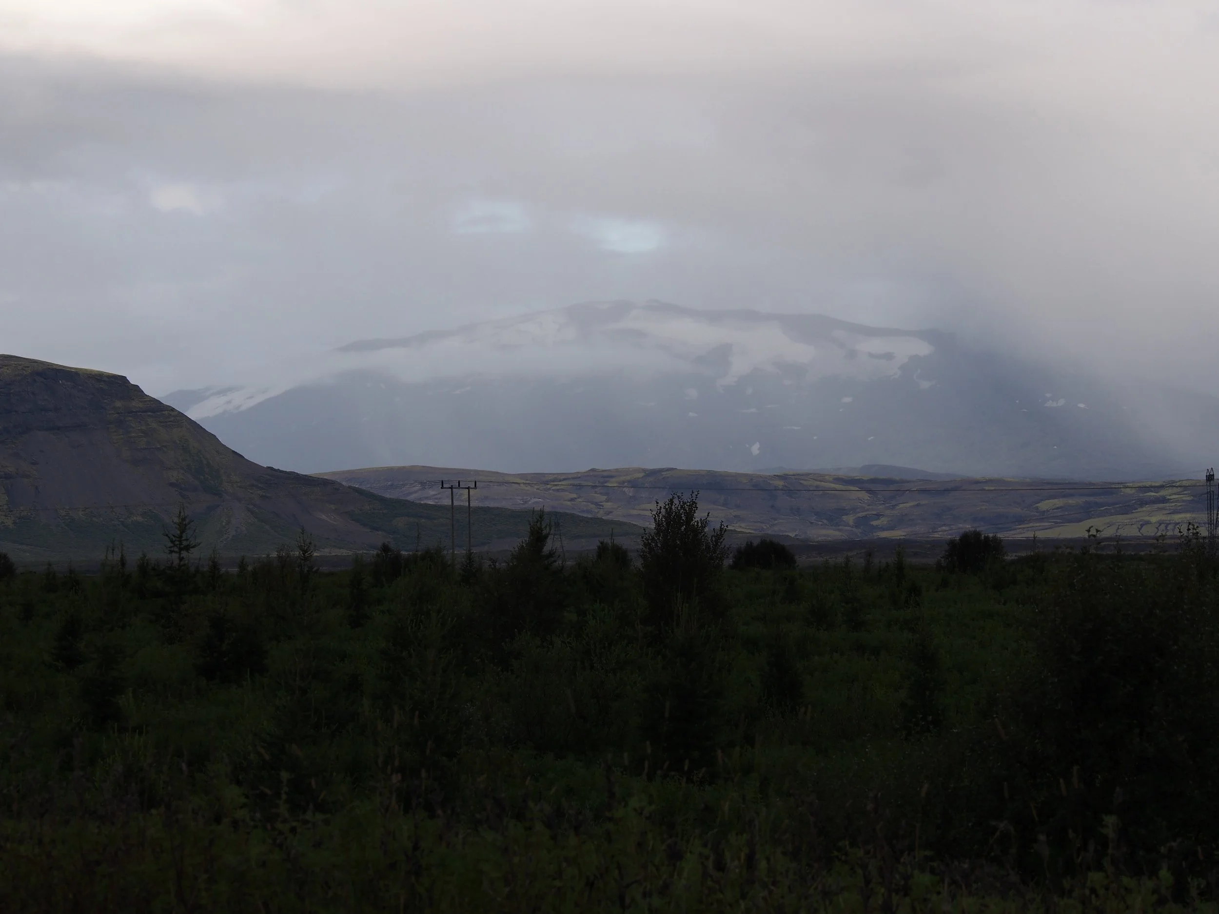 A landscape with mountains (Hekla) in the distance, partly covered with snow, under a cloudy sky; foreground consists of dark green vegetation and trees.