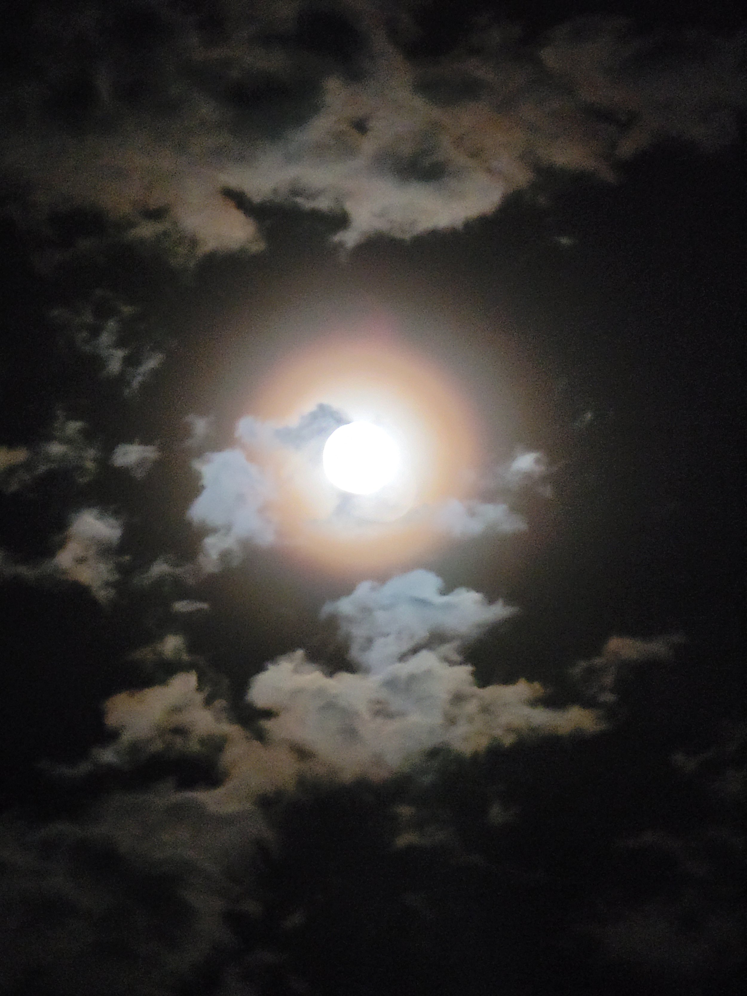 An image of the full moon with corona with clouds floating past. Almost like a painting.