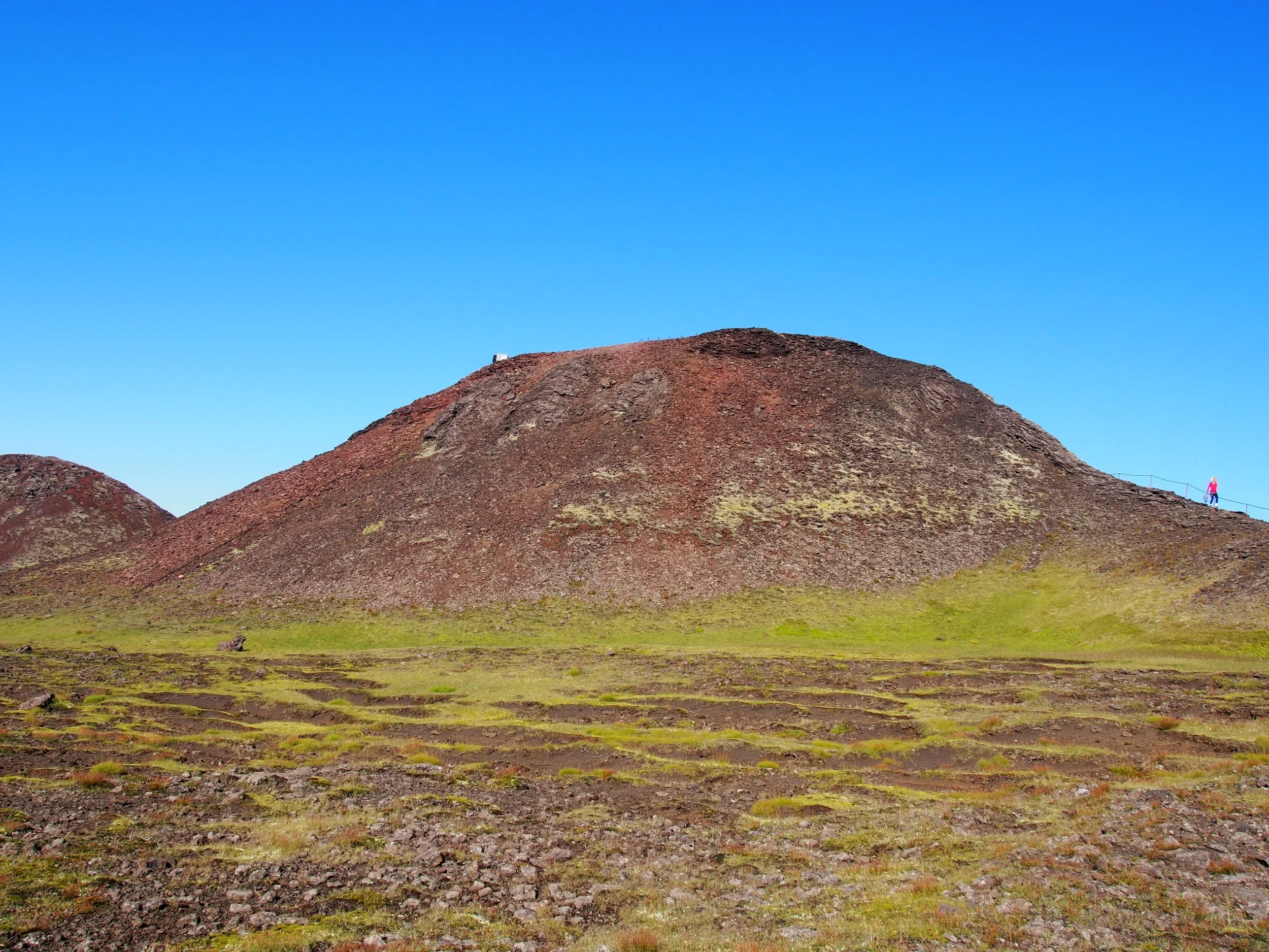 Approaching Þríhnúkagígur with a green foreground and a clear blue sky. A person in red is walking along a path on the right side of the hill.