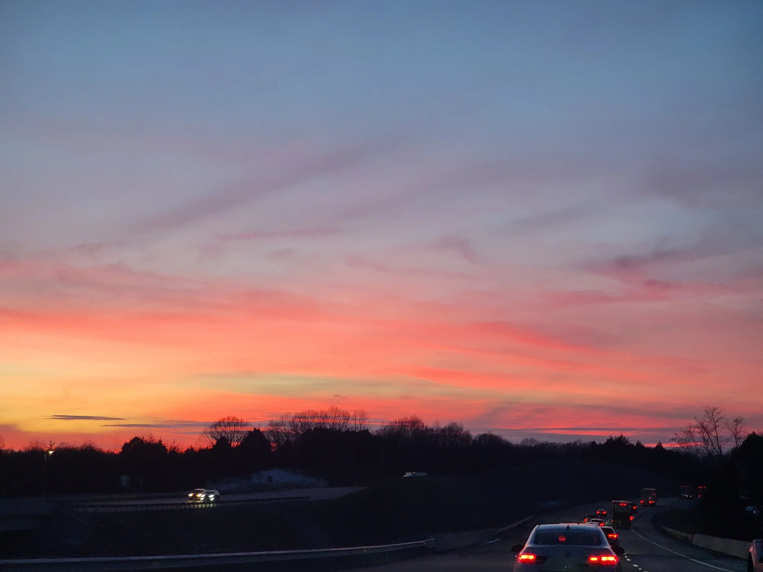 A highway during sunset with pink and purple clouds in the sky and several cars traveling on the road.