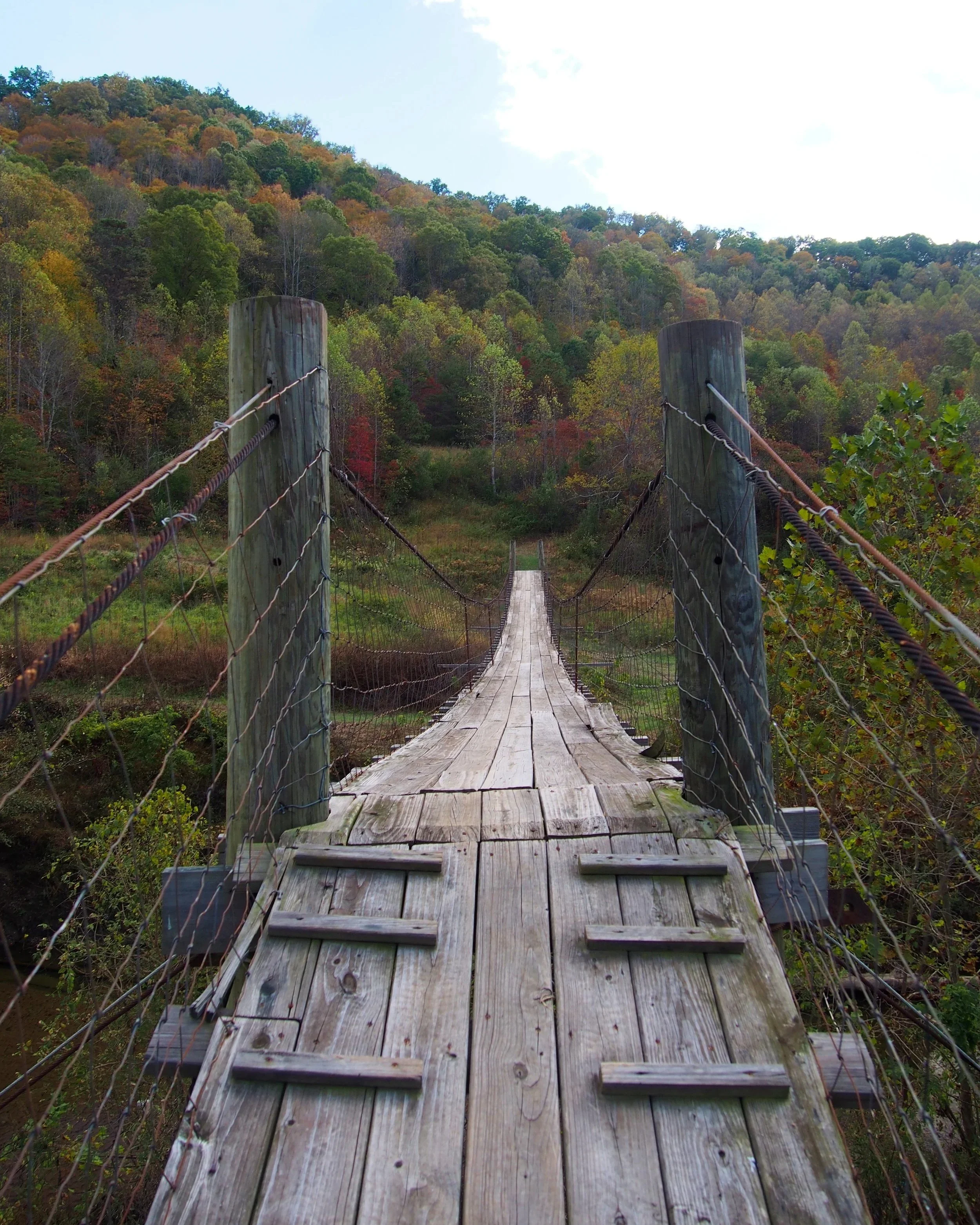 A wooden suspension bridge extending over a lush, autumn landscape with colorful trees and a forested hill in the background.