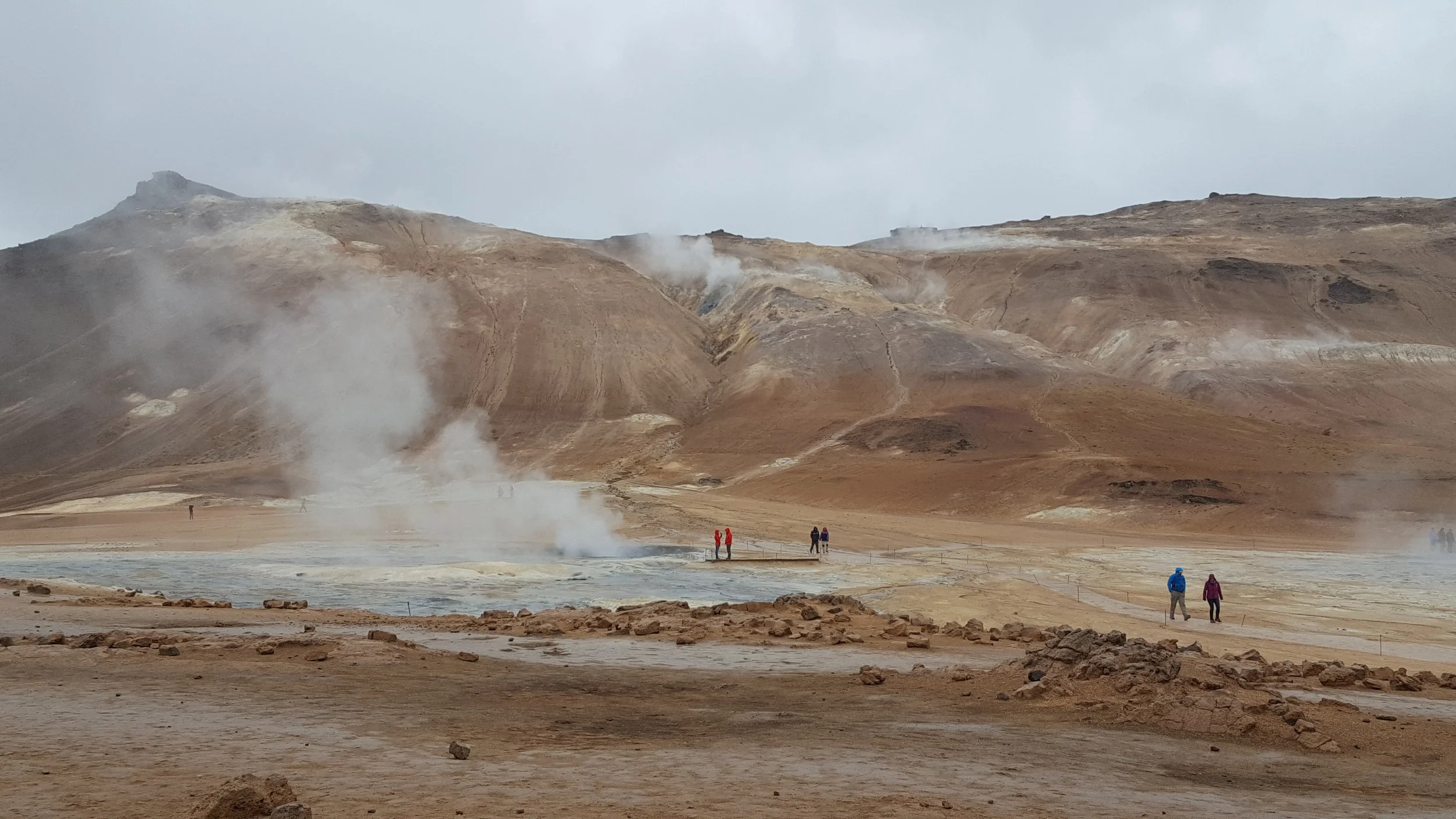 A volcanic area with steam vents, barren rocky terrain, and hills in the background, with a few people walking around in outdoor clothing.