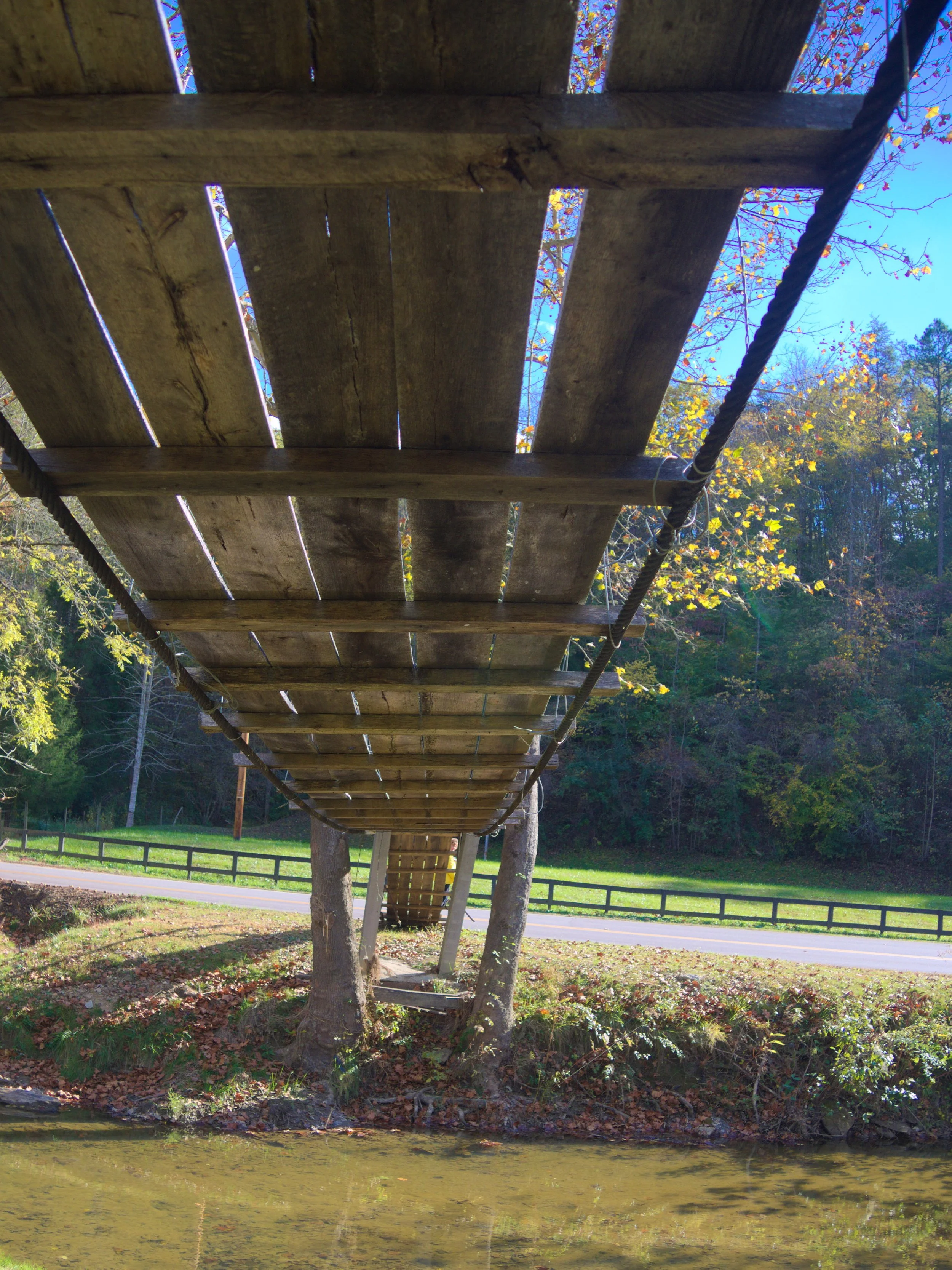 Underneath view of a wooden footbridge over a creek with trees and a grassy area in the background.