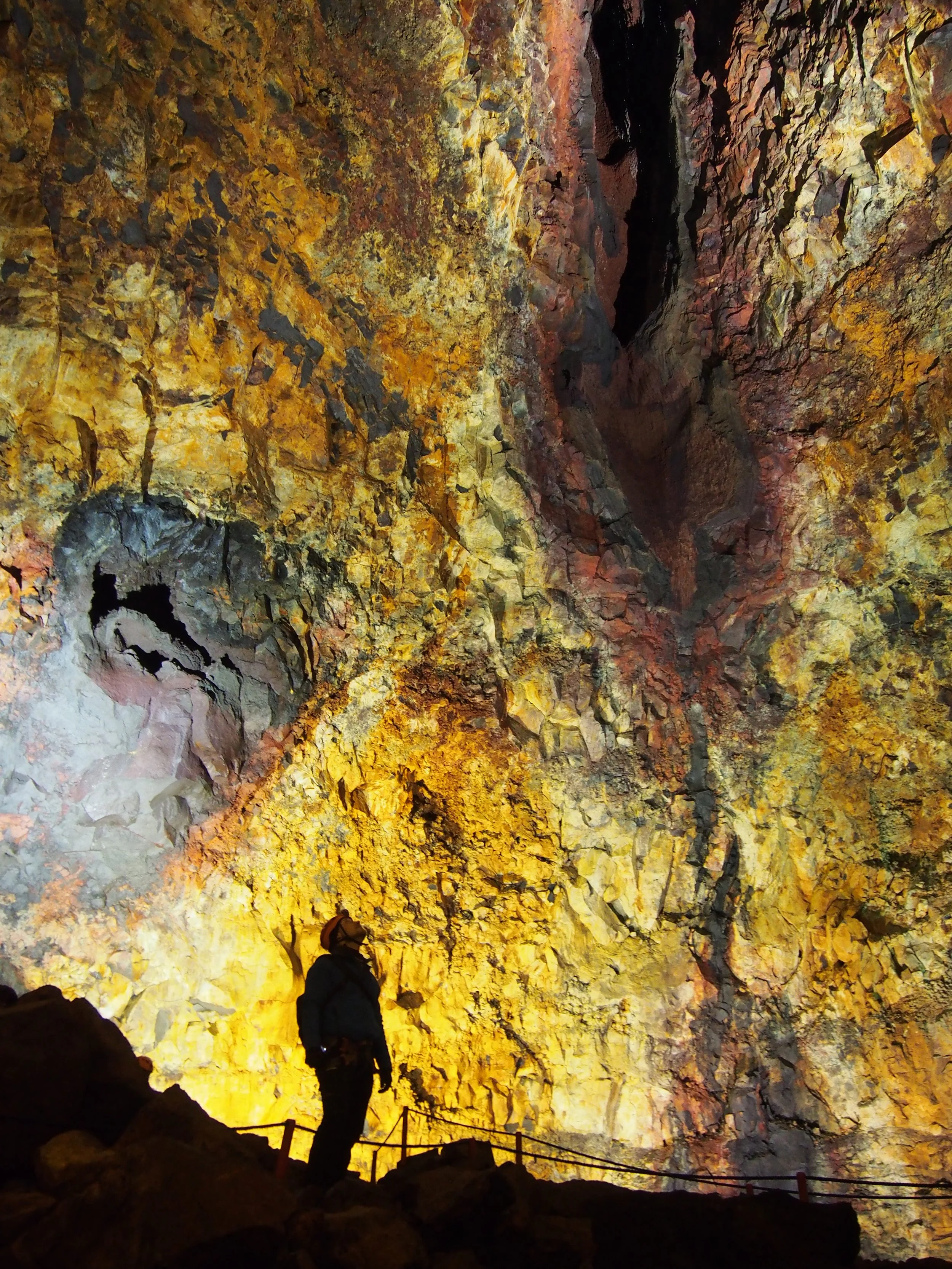 Person standing inside a large, colorful cave with yellow, orange, and red rock formations illuminated by lights.