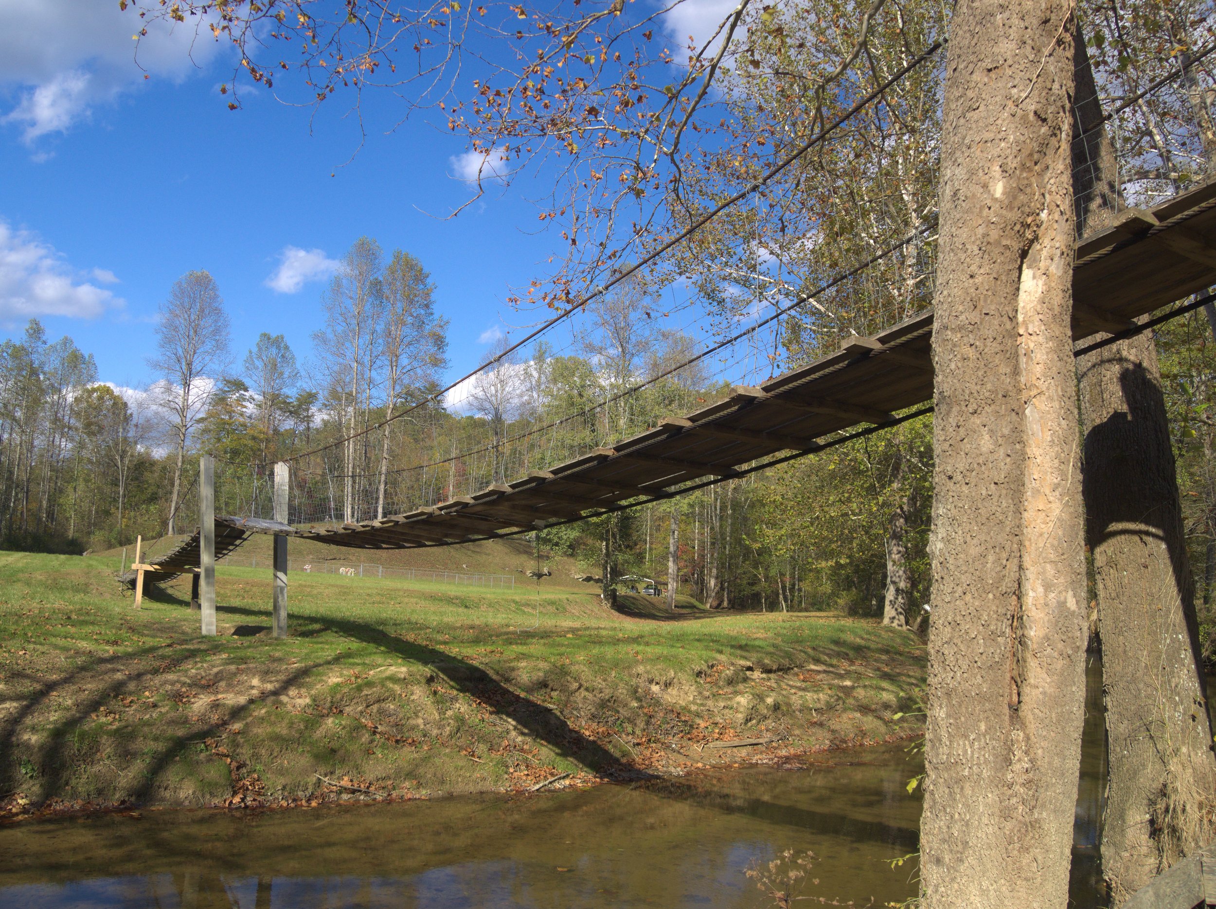 A suspension bridge over a creek in a wooded park with trees, grass, and blue sky with some clouds.