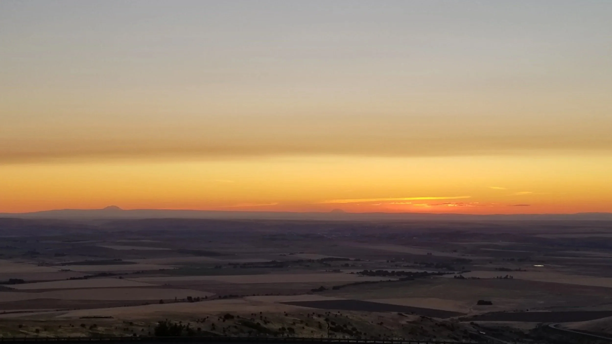 Sunset over a wide, flat landscape with distant mountains on the horizon and a colorful sky with orange and yellow hues.