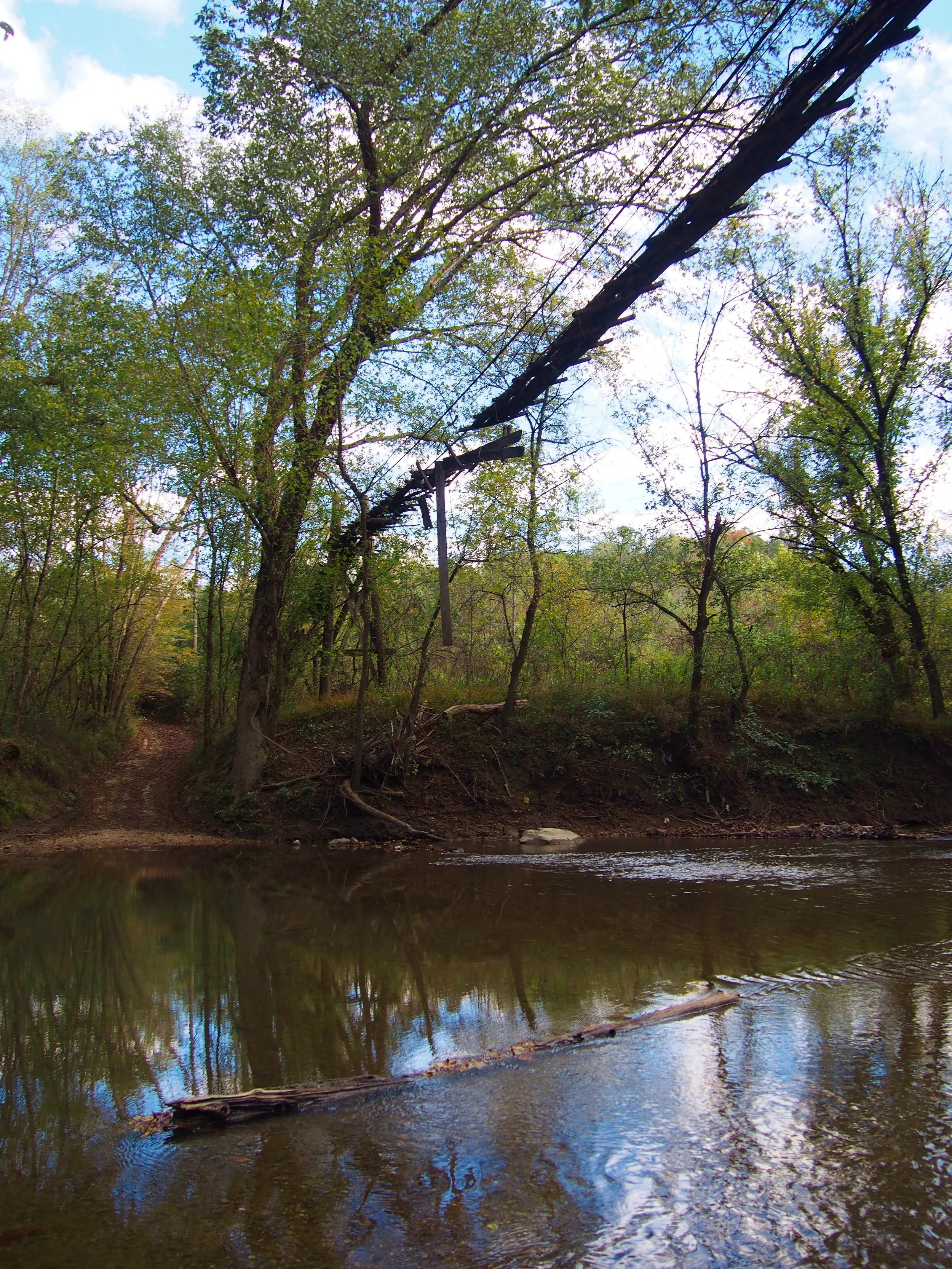 Overhanging, derelict bridge above a small river in a forest with green trees and a dirt path on the left.