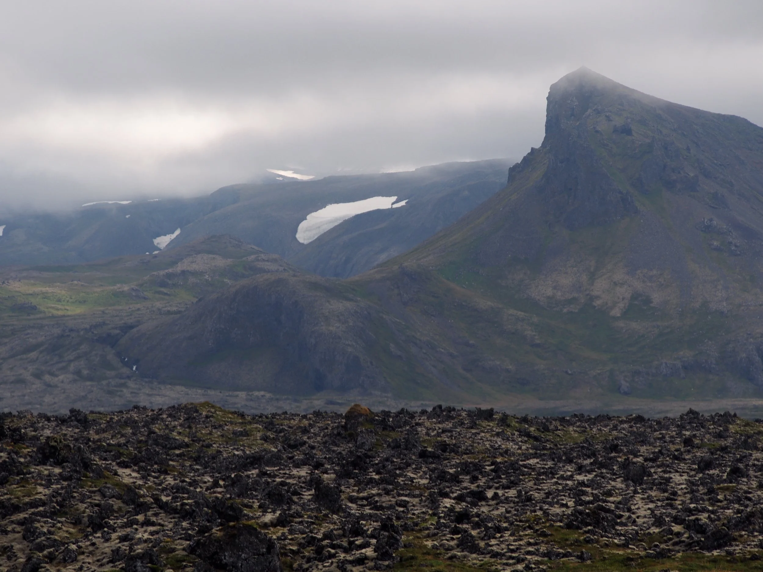 A rugged mountain (Snæfellsjökull) landscape with a prominent steep mountain and patches of snow, under a cloudy sky.