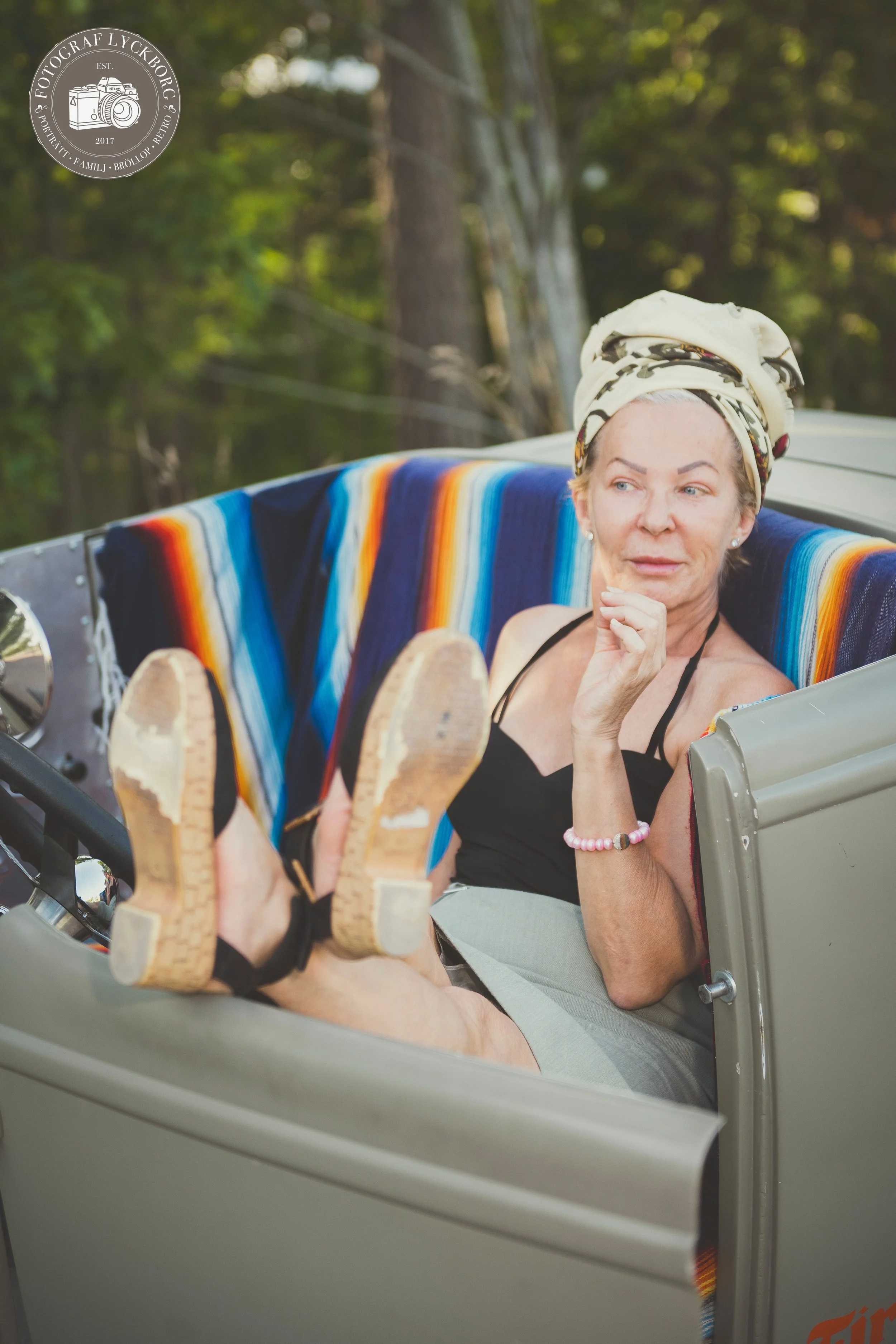 En äldre kvinna som sitter i ett fordon, på sig har hon ett mönstrat huvudband, svart tröja och ljusgrå kjol, hon vilar hakan på handen och tittar åt sidan, sittandes på en färgglad, randig filt draperad över ryggen.