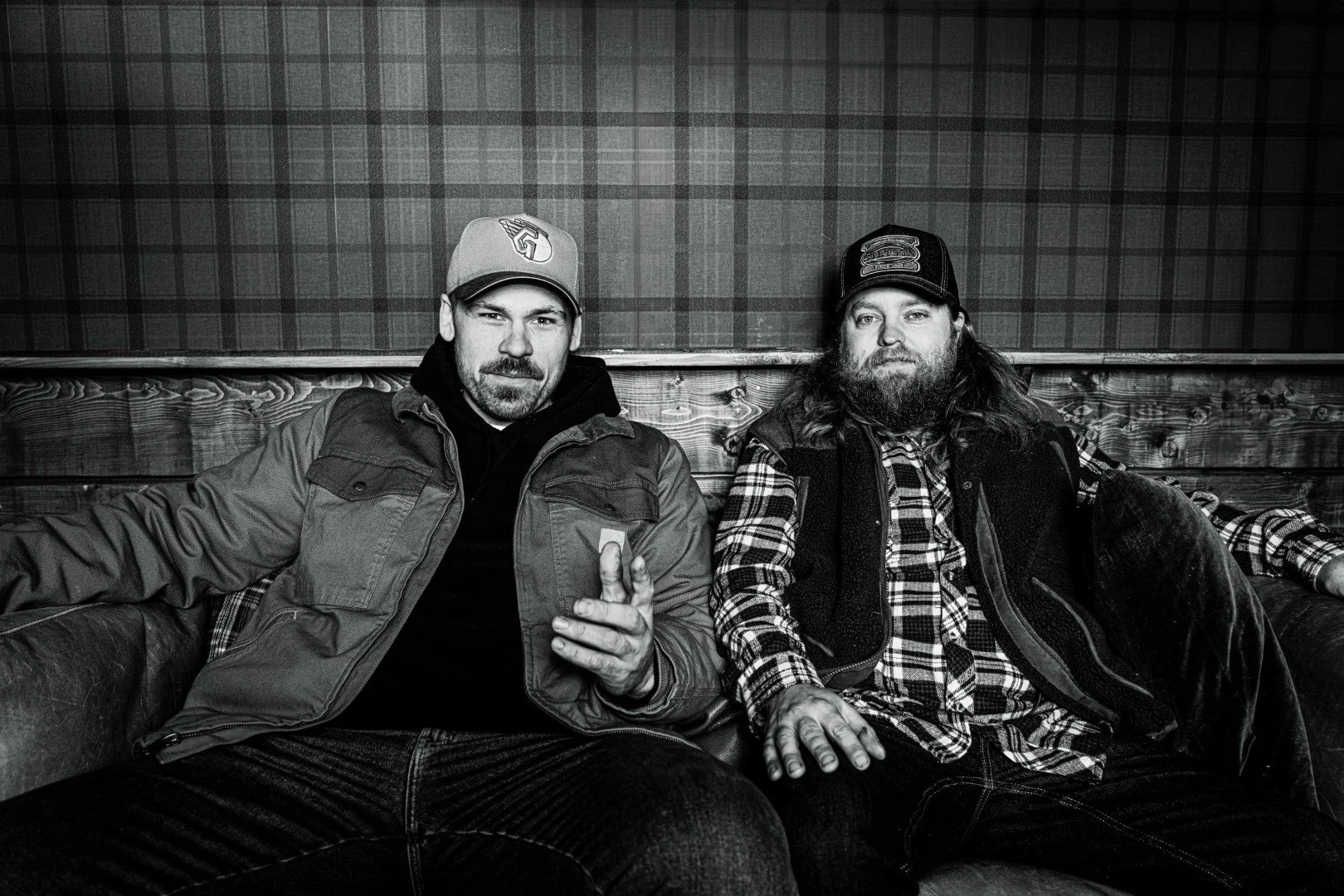 Två män sitter på en svart skinnsoffa i ett rum med träpanelväggar och en tapet med rutmönster. Den vänstra mannen har en keps och en jacka, den högra mannen har en keps och en flanellskjorta. Båda tittar mot kameran.