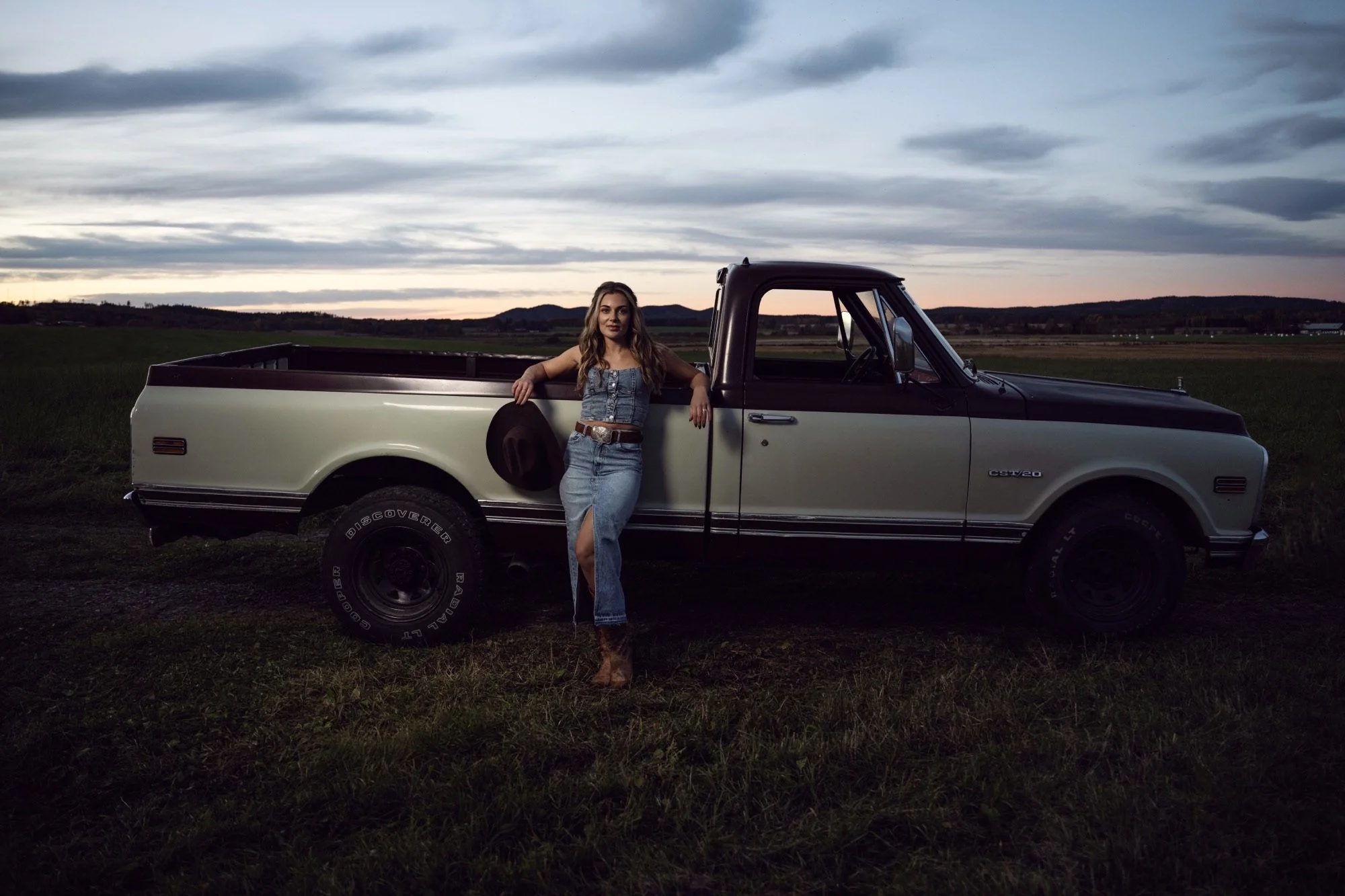 En ung kvinna står bredvid en vit och svart äldre pickup-truck i ett öppet landskap vid solnedgången.