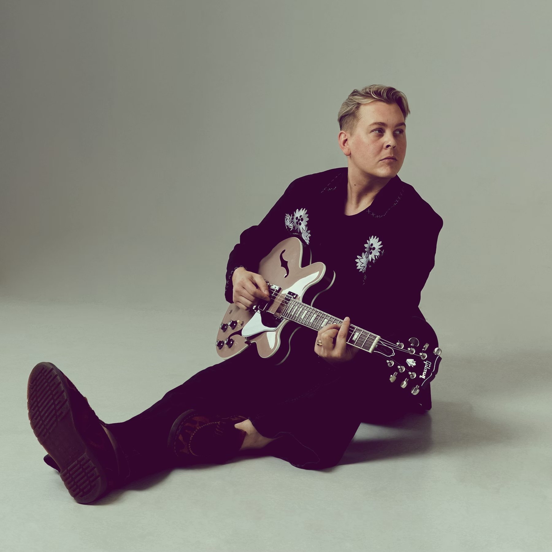 En person sitter på golvet och spelar gitarr, klädd i svart med blommönster på jackan, mot en neutral bakgrund.