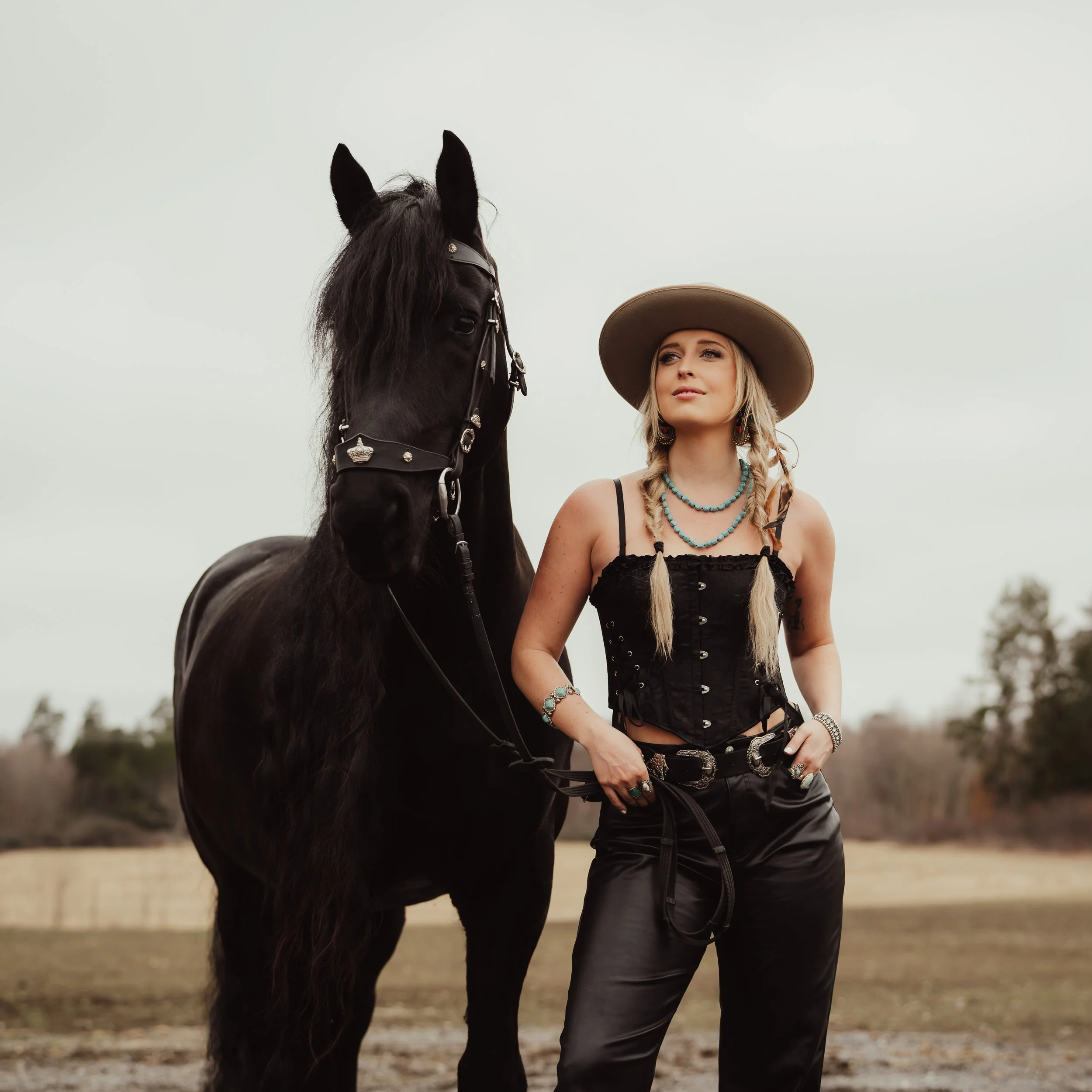 En kvinna står bredvid en svart häst i ett öppet landskap, både ser ut att vara fotograferade i en landsflykt.